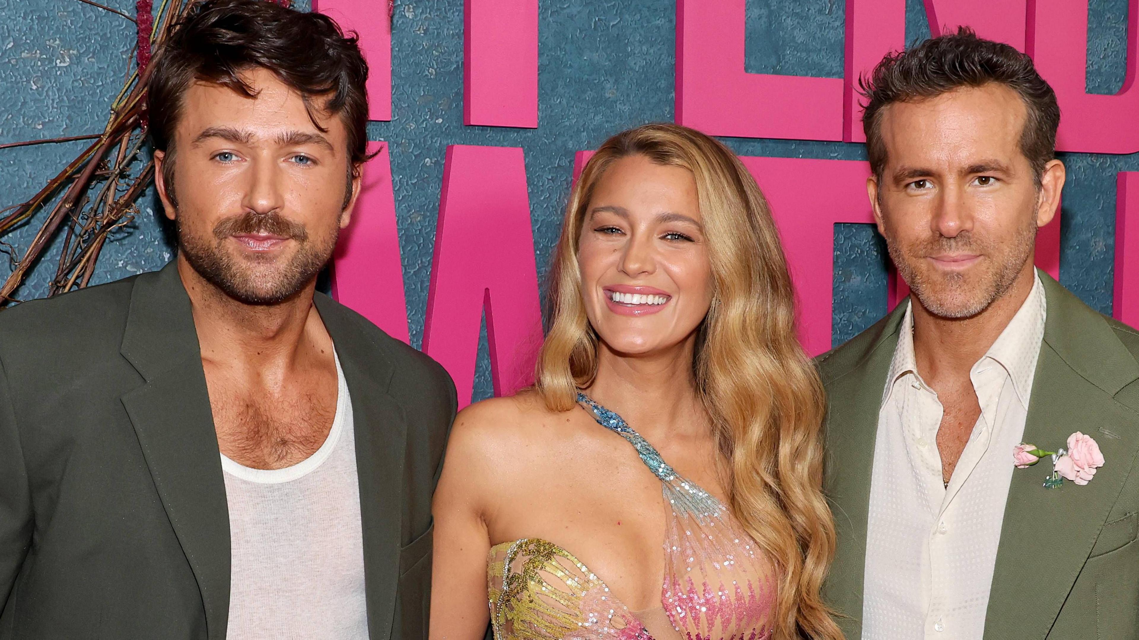 Three people - Brandon Sklenar, Blake Lively and Ryan Reynolds - are dressed in formal attire and posing for a photo. They are stood in front of a large sign that reads "It Ends With Us"