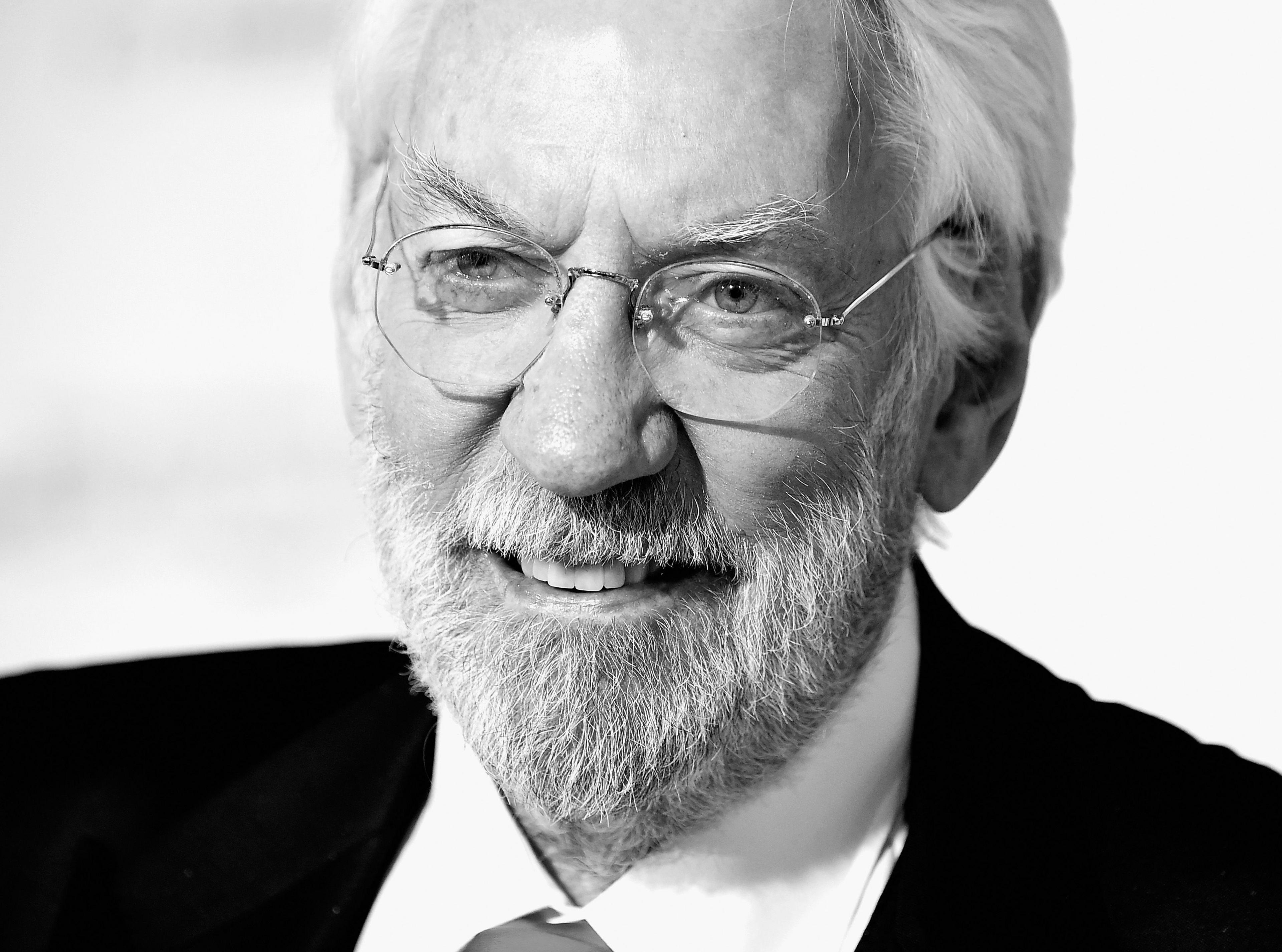 Donald Sutherland in a black-and-white image, wearing wire-rimmed glasses - he has white hair and a grey/white beard and wears a black suit 