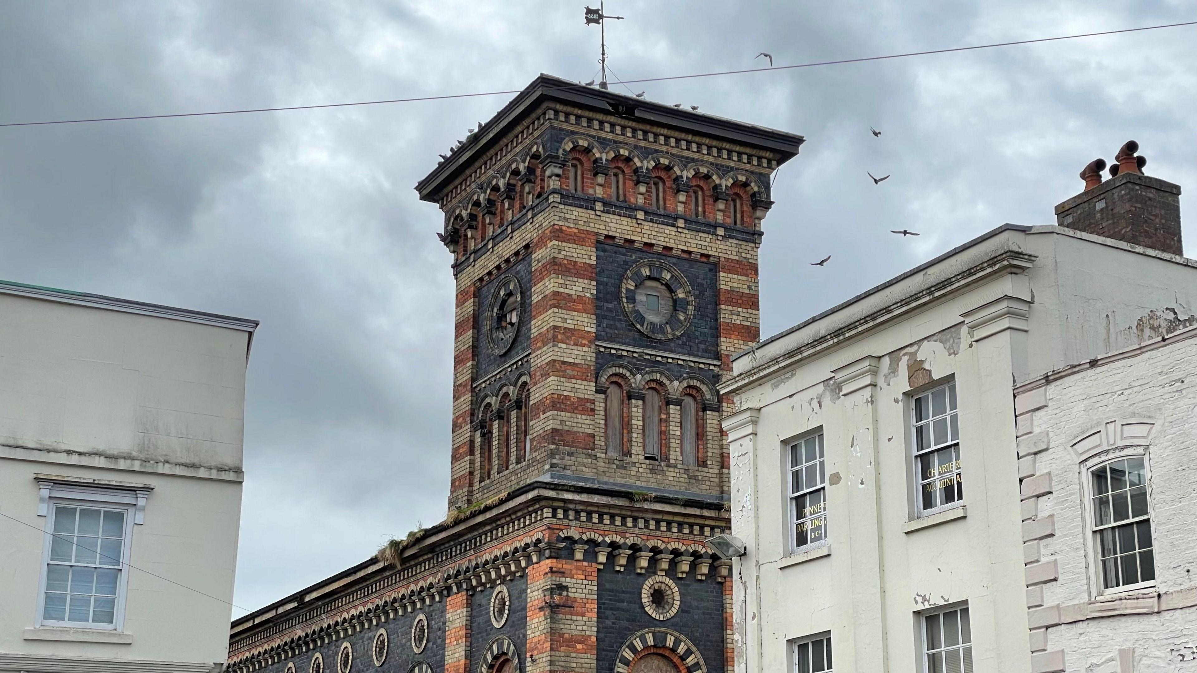 The New Market Buildings, which is of Italianate architecture, with pigeons flying around it's tower, which is topped with a weather vane