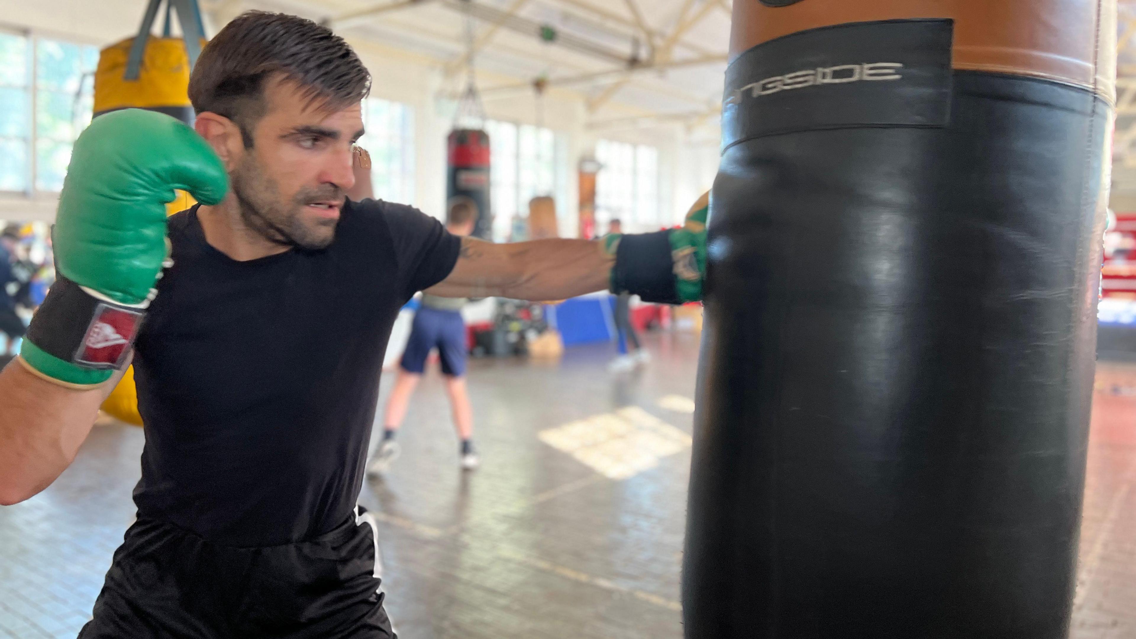 Chris punches a heavy punching bag that's hanging from the ceiling as people train behind him in the boxing club