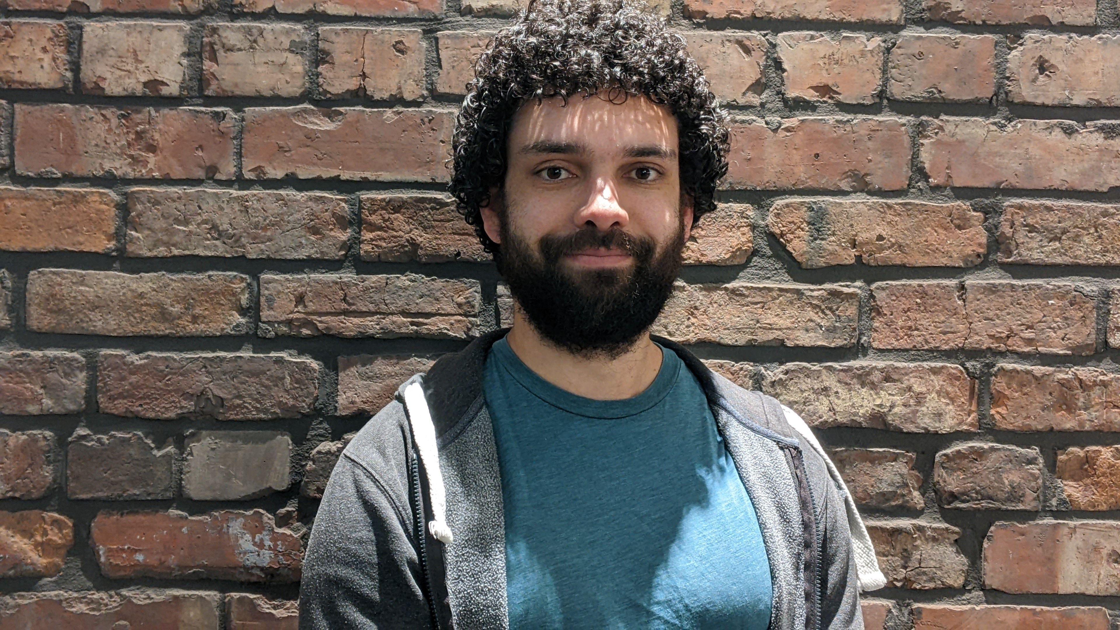 Nick Weise stands against a weathered brick wall. He has curly dark hair and a short beard, and is wearing a teal T‑shirt under a dark grey zip‑up hoodie. 