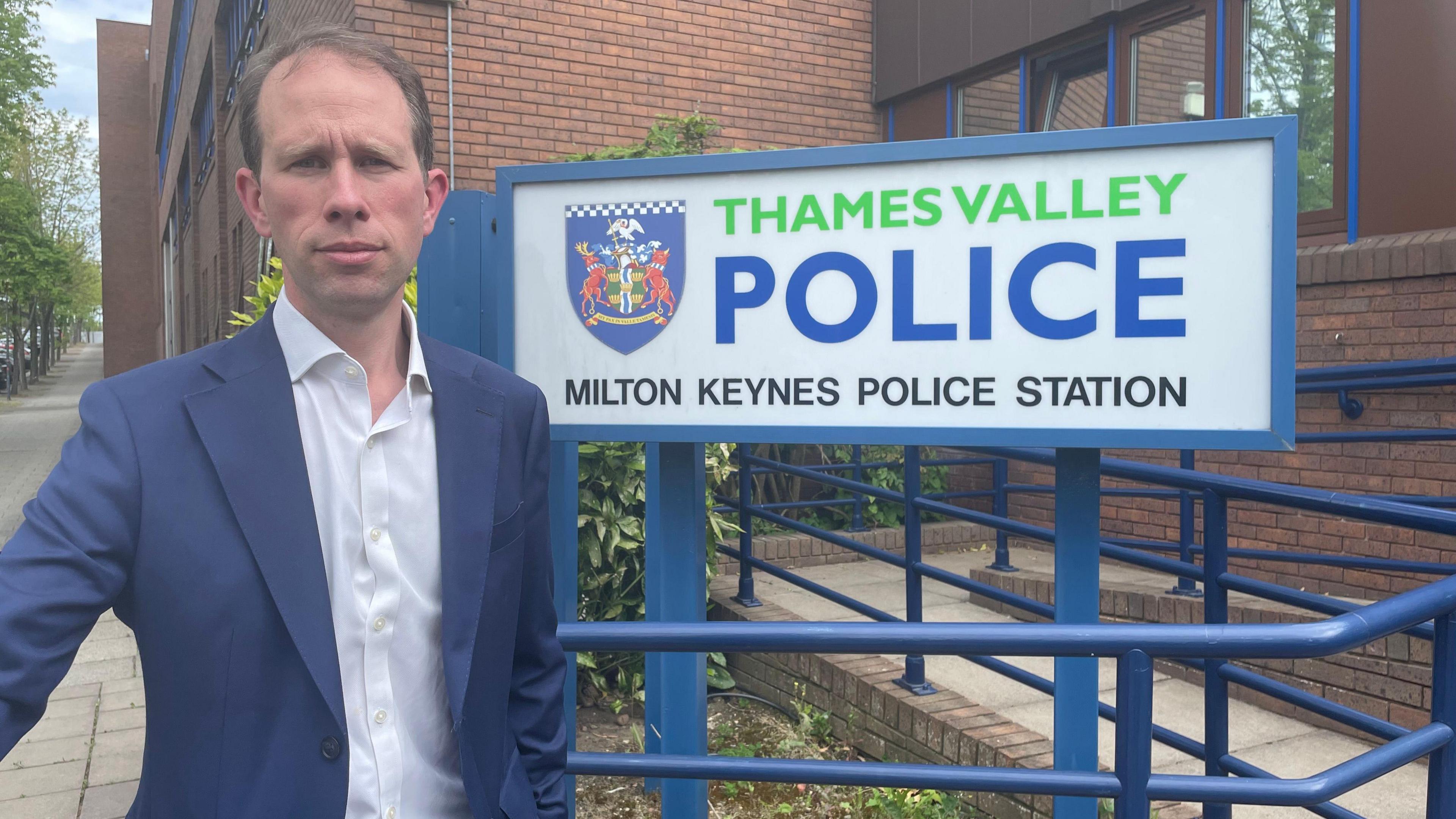 Matthew Barber has short brown hair and is wearing a white shirt and blue blazer. He is stood in front of Thames Valley Police's Milton Keynes station.