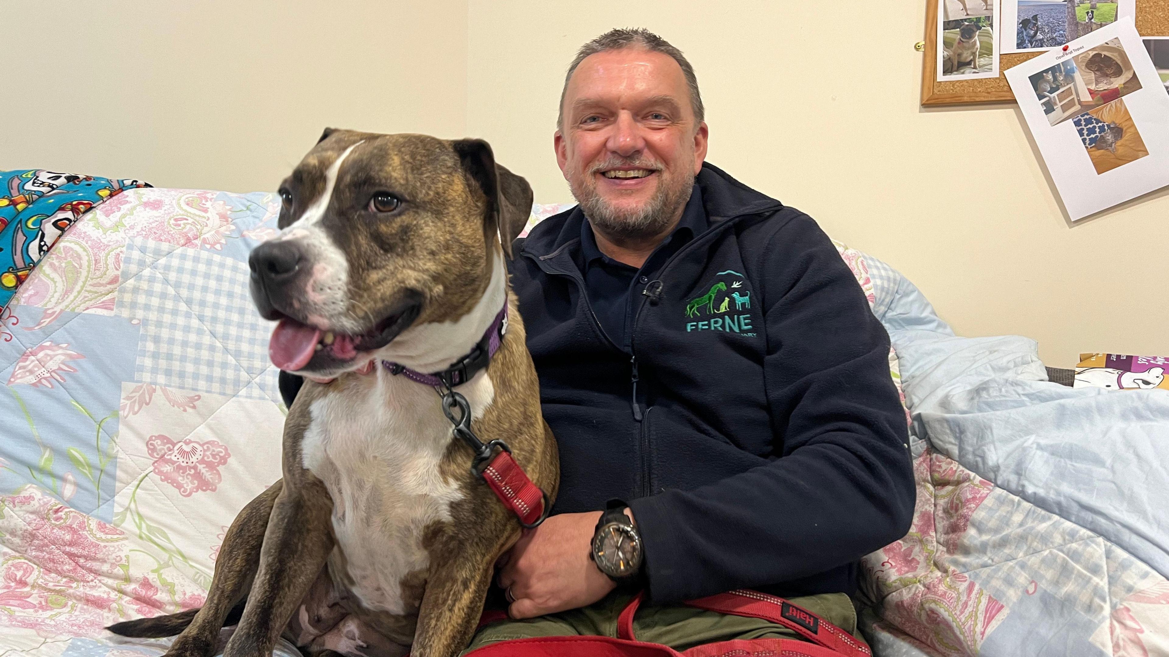 Kevan Hodges wearing a Ferne Animal Sanctuary dark blue zip jacket, and sitting on a sofa with a large brown and white dog