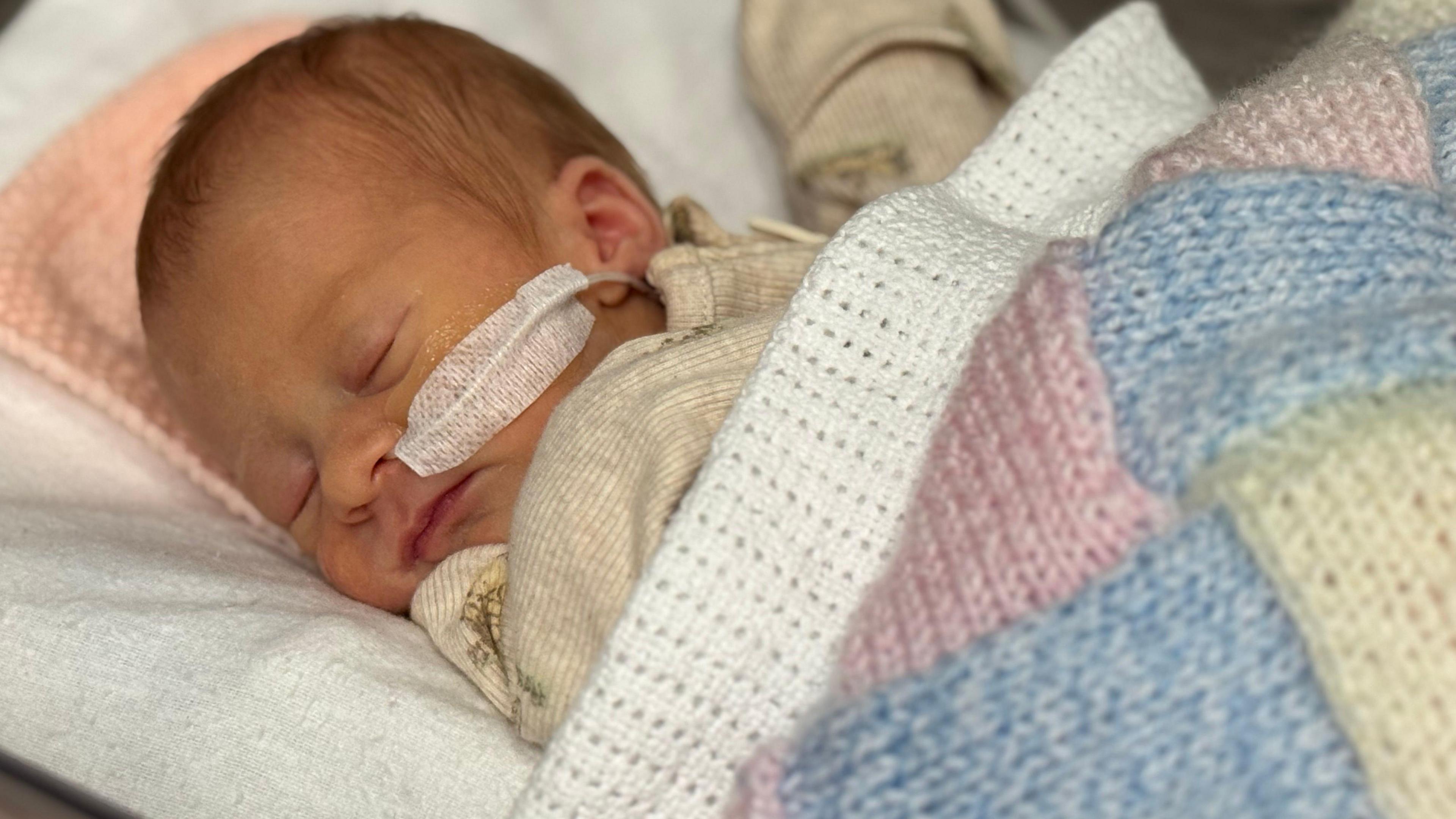 A baby is lying on her side asleep in a hospital bed, while facing the camera. The baby has a tube up her nose and is resting her head on a pillow. She has been covered in a multi-coloured blanket.