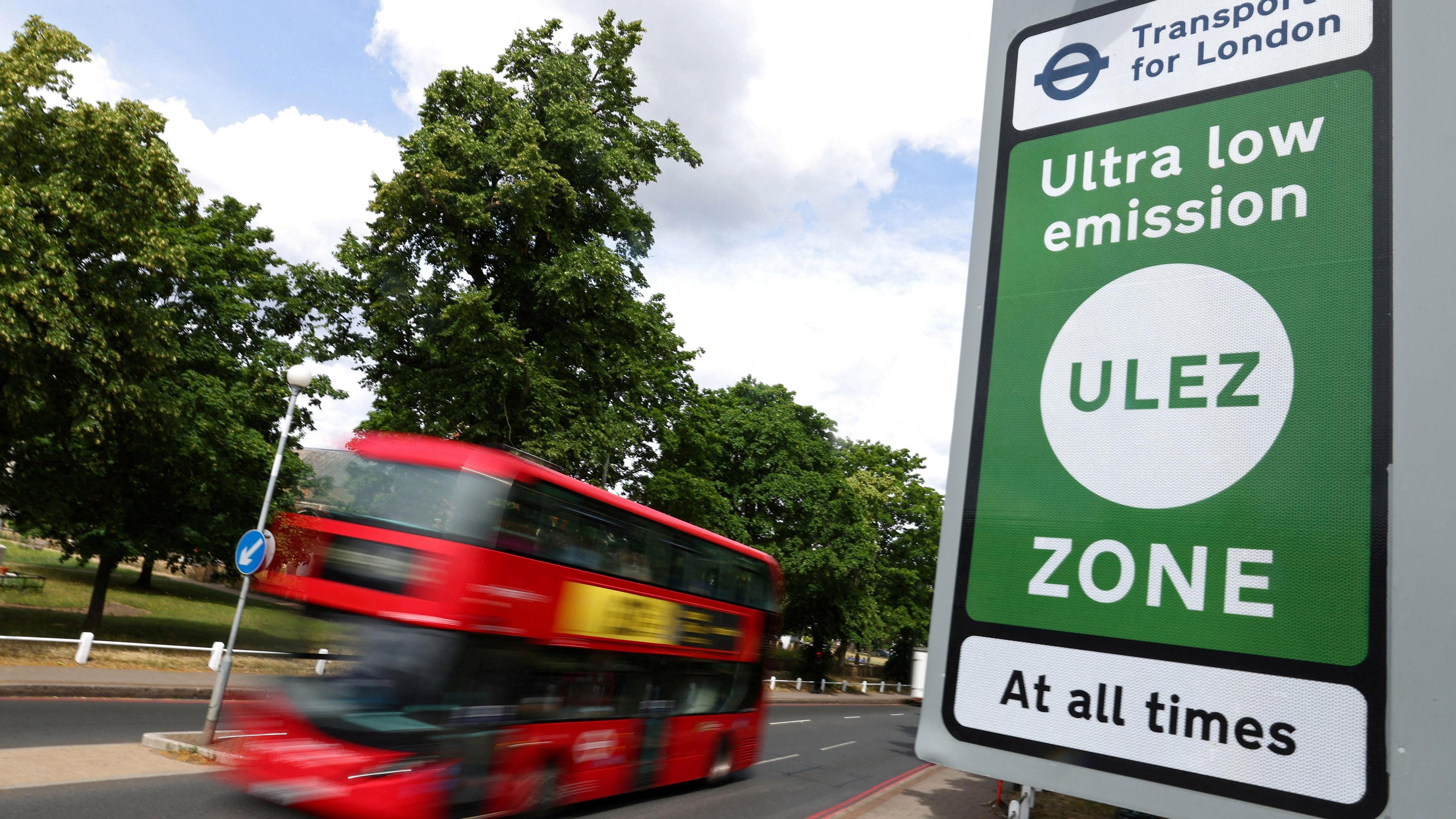 A blurry red double-decker bus drives past a green TfL sign for the Ultra Low Emission Zone (Ulez)