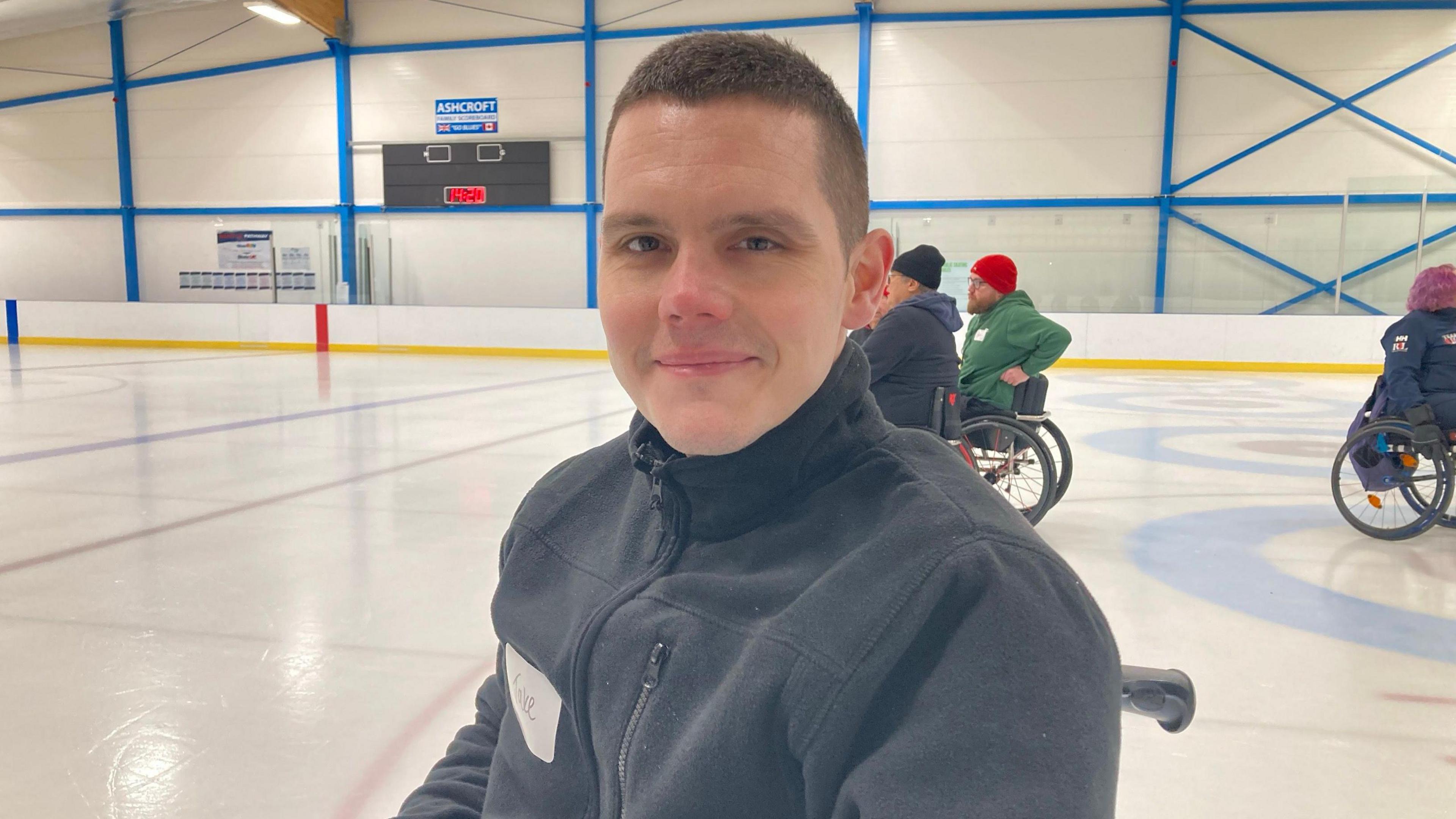 Jake McHale is looking at the camera and smiling. He is on the ice in his chair, wearing a grey jacket. People in wheelchairs can be seen on the rink behind him