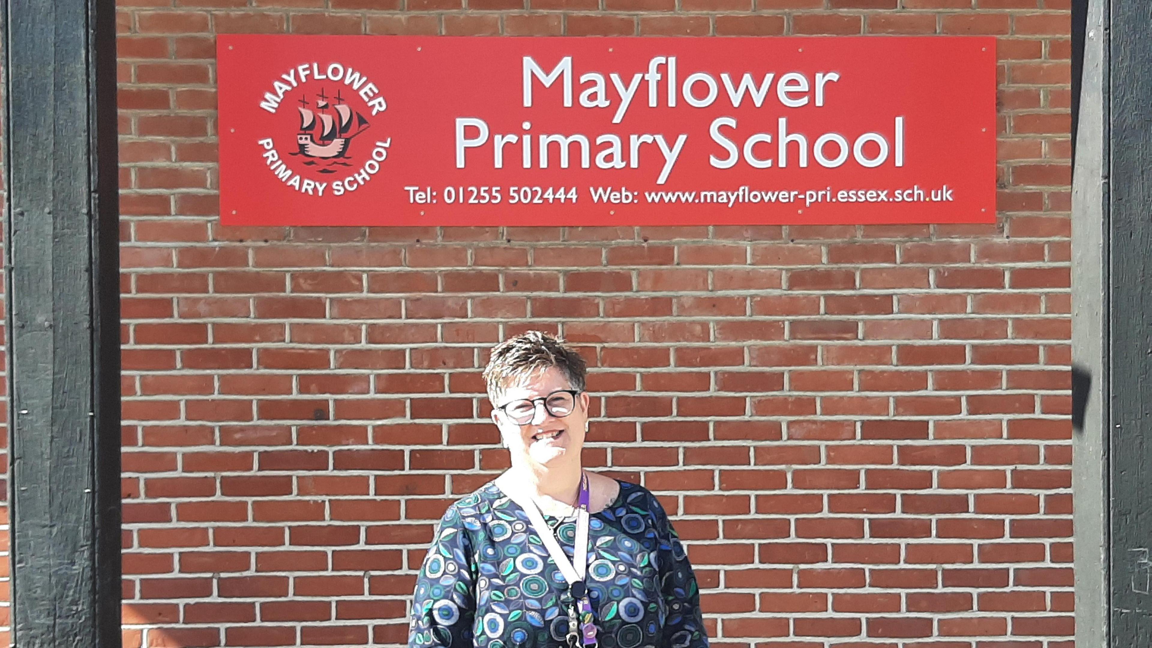 A female head teacher with short hair standing and smiling in front of her school's sign, which is on a brick wall.
