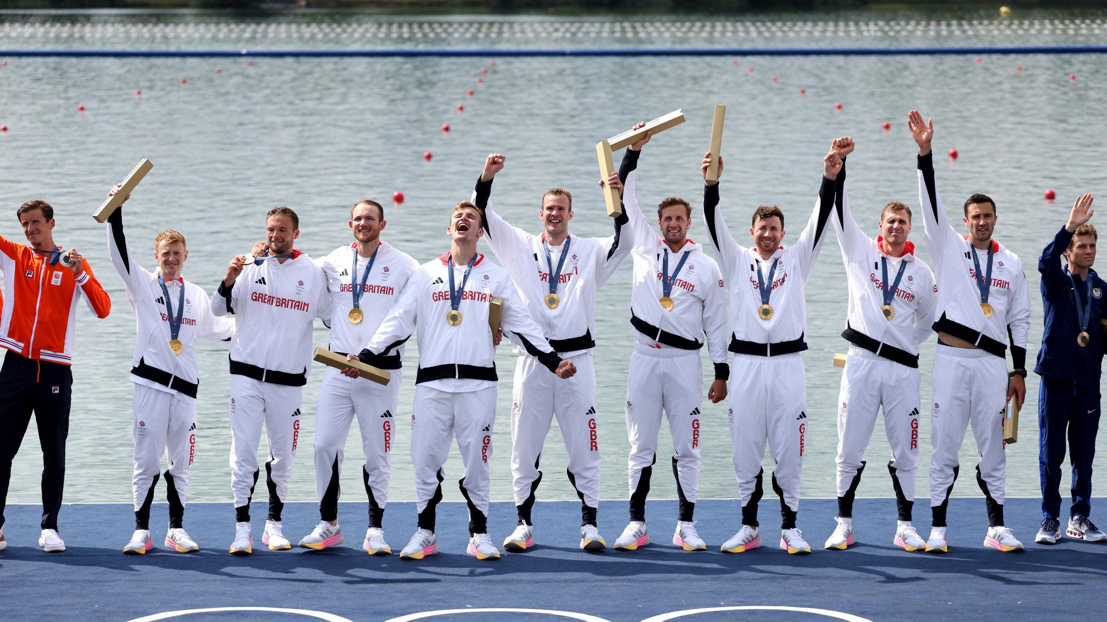 The victorious GB Eights team standing on the medal podium at the Paris Olympics in August 2024. They wear white GB kit and all have medals around their necks