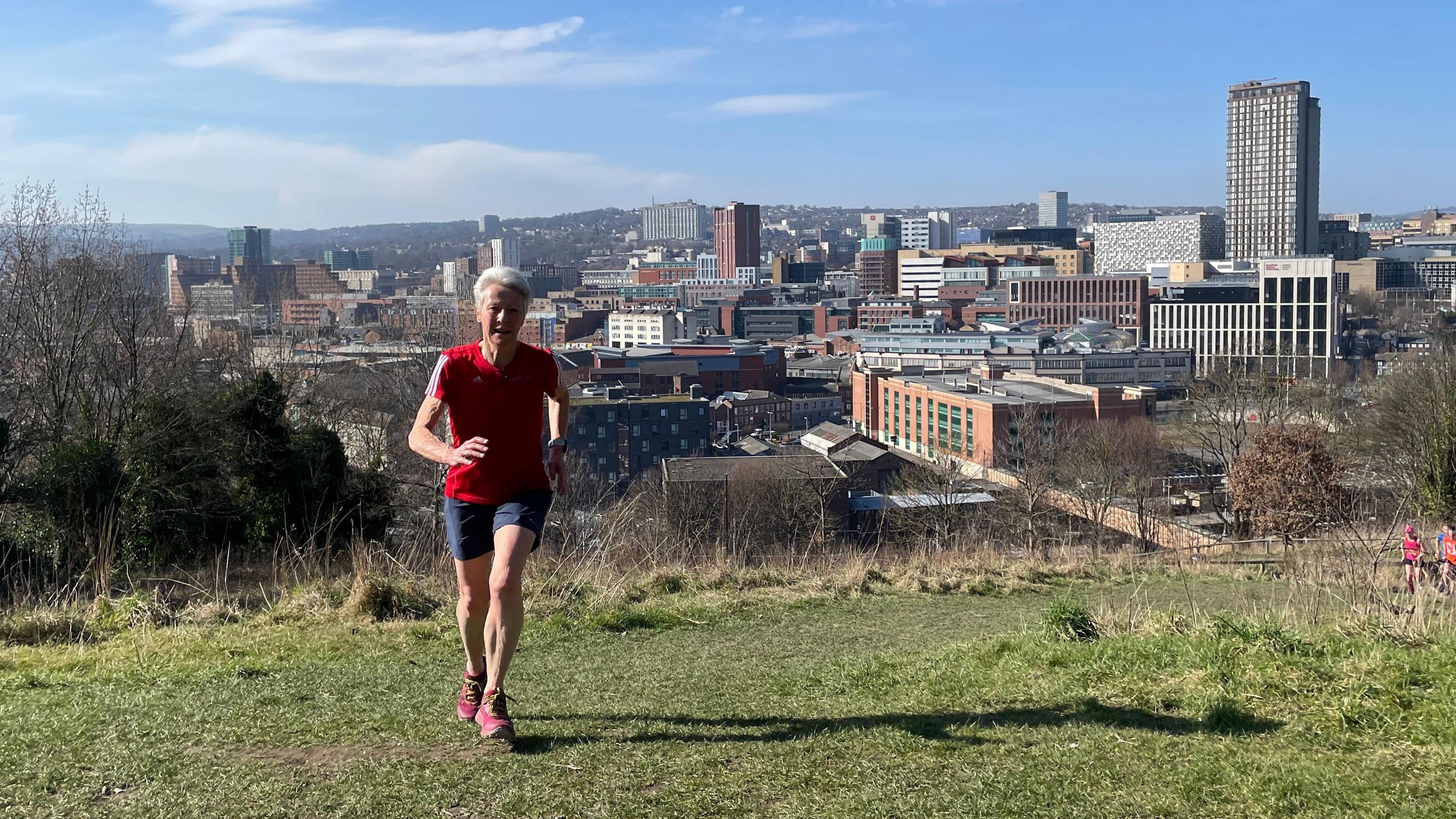 Dot Kesterton running in Clay Wood, Sheffield, where she trains with the backdrop of the city centre behind her. She's wearing a red Parkrun t shirt, blue shorts and red trainers. It's a sunny, Spring day.