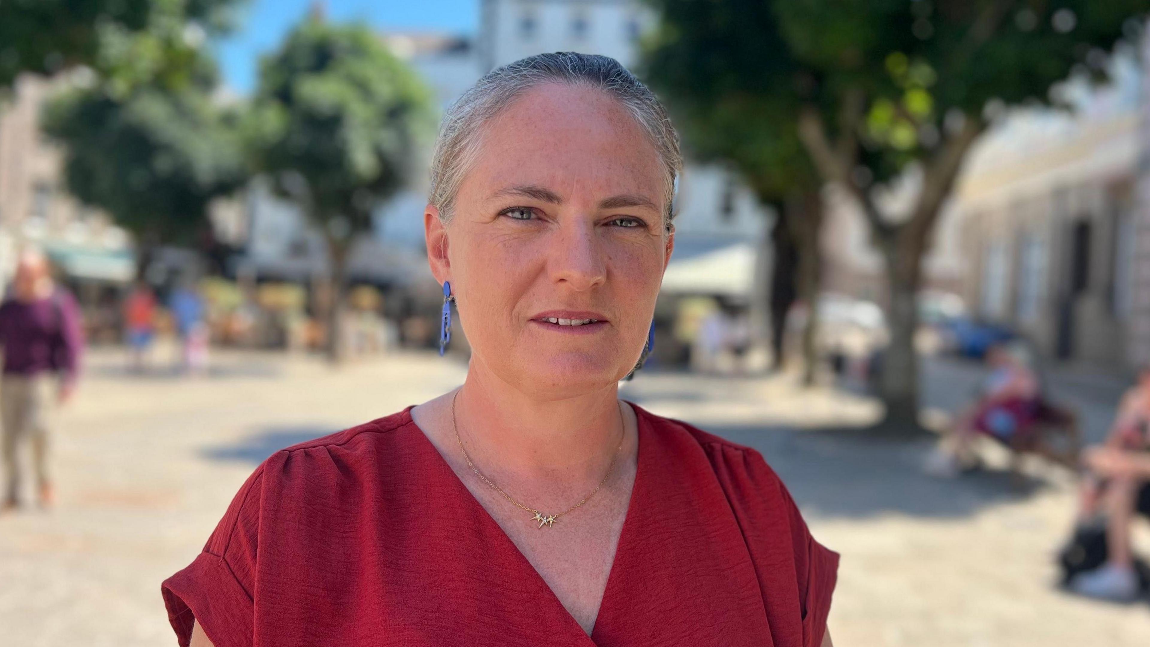 Deputy Hilary Jeune is standing in Jersey's Royal square, she's wearing a red top with a V-neck and blue earrings.
