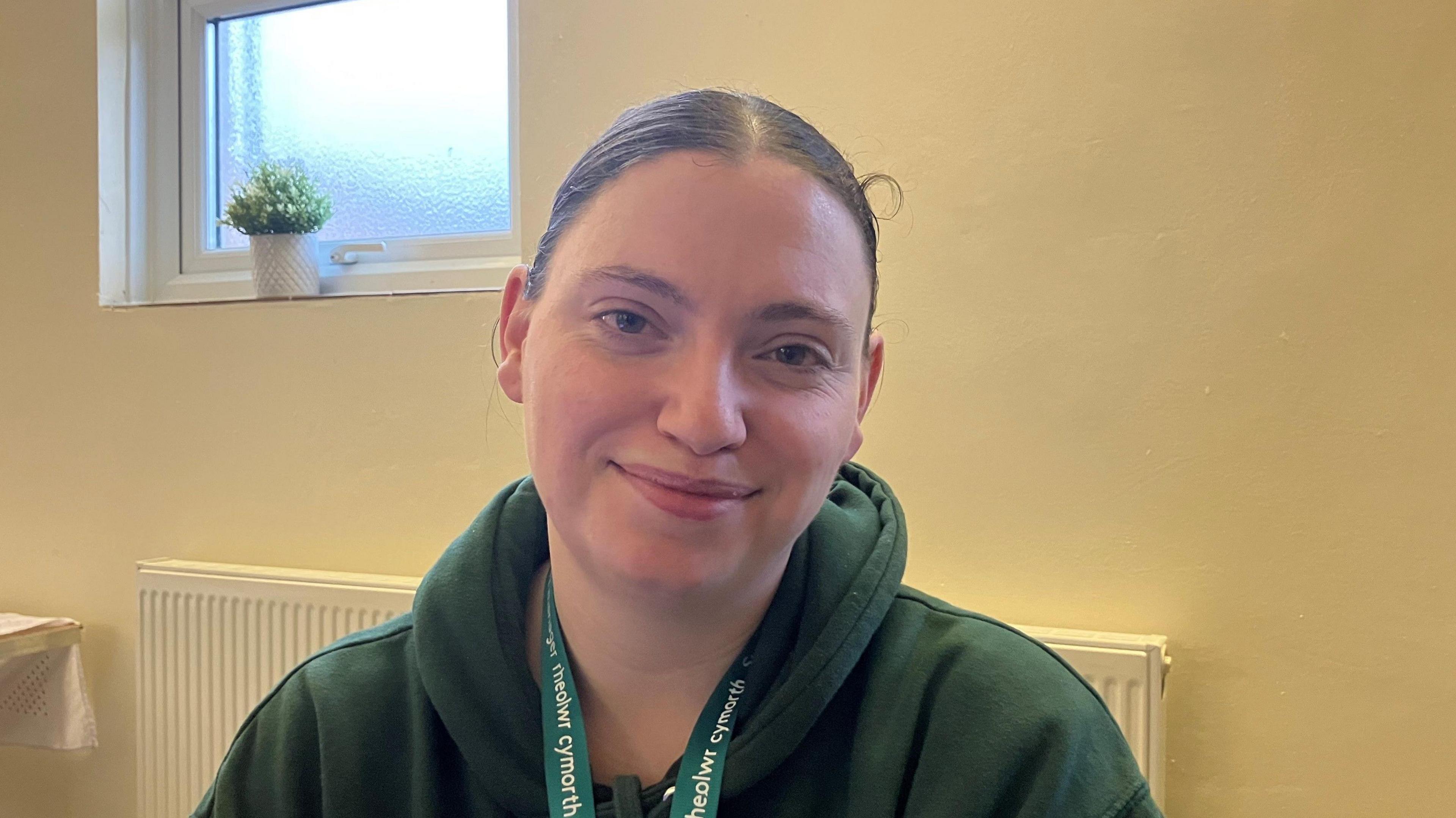 A young woman with brown hair tied back, wearing a green hoodie and sitting at a desk at a Taff Ely Foodbank site. 