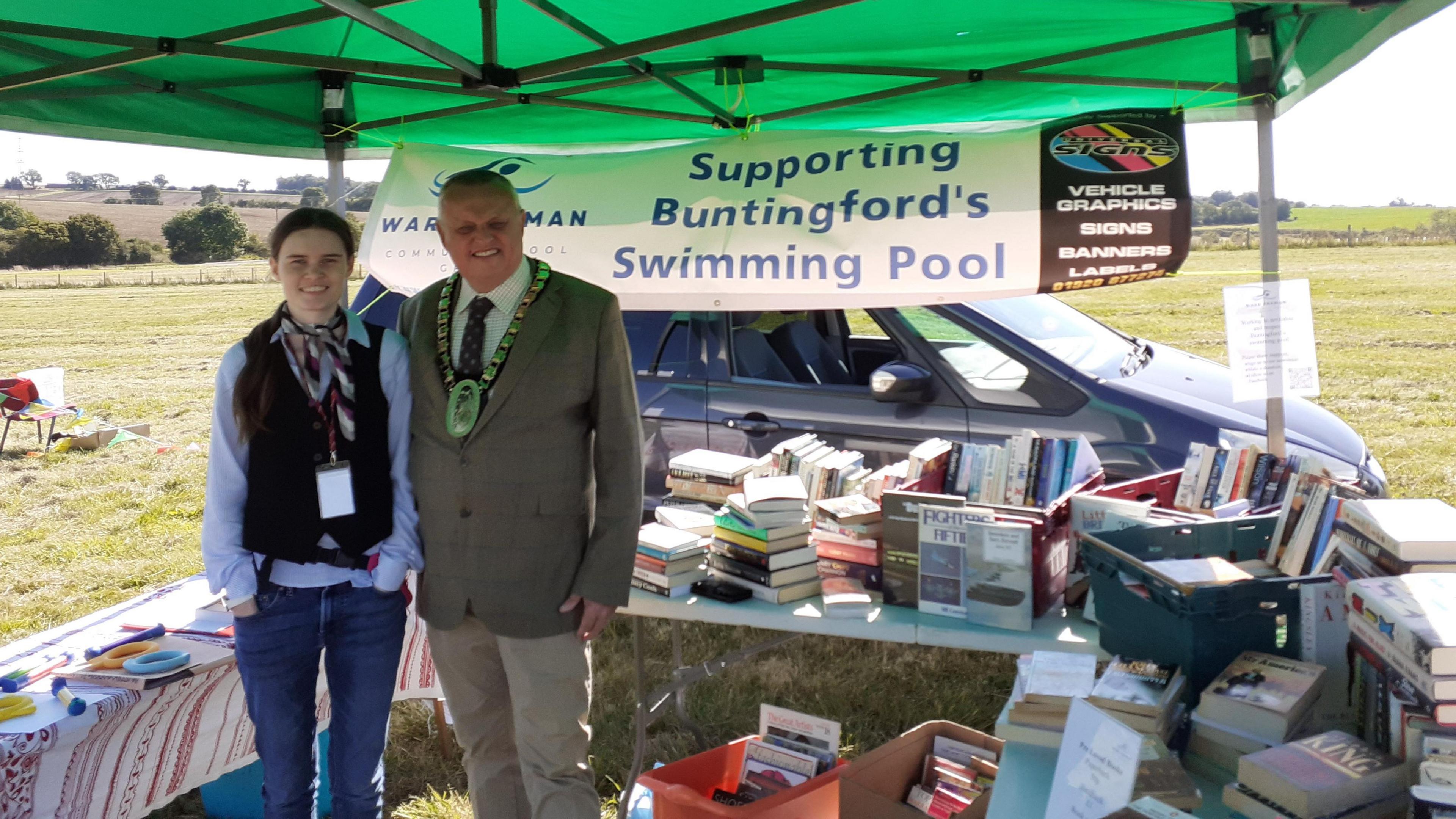 A man, wearing a mayoral chain, and a jacket, standing next to a young woman, at a book stall, by a banner that says "Supporting Buntingford's Swimming Pool"