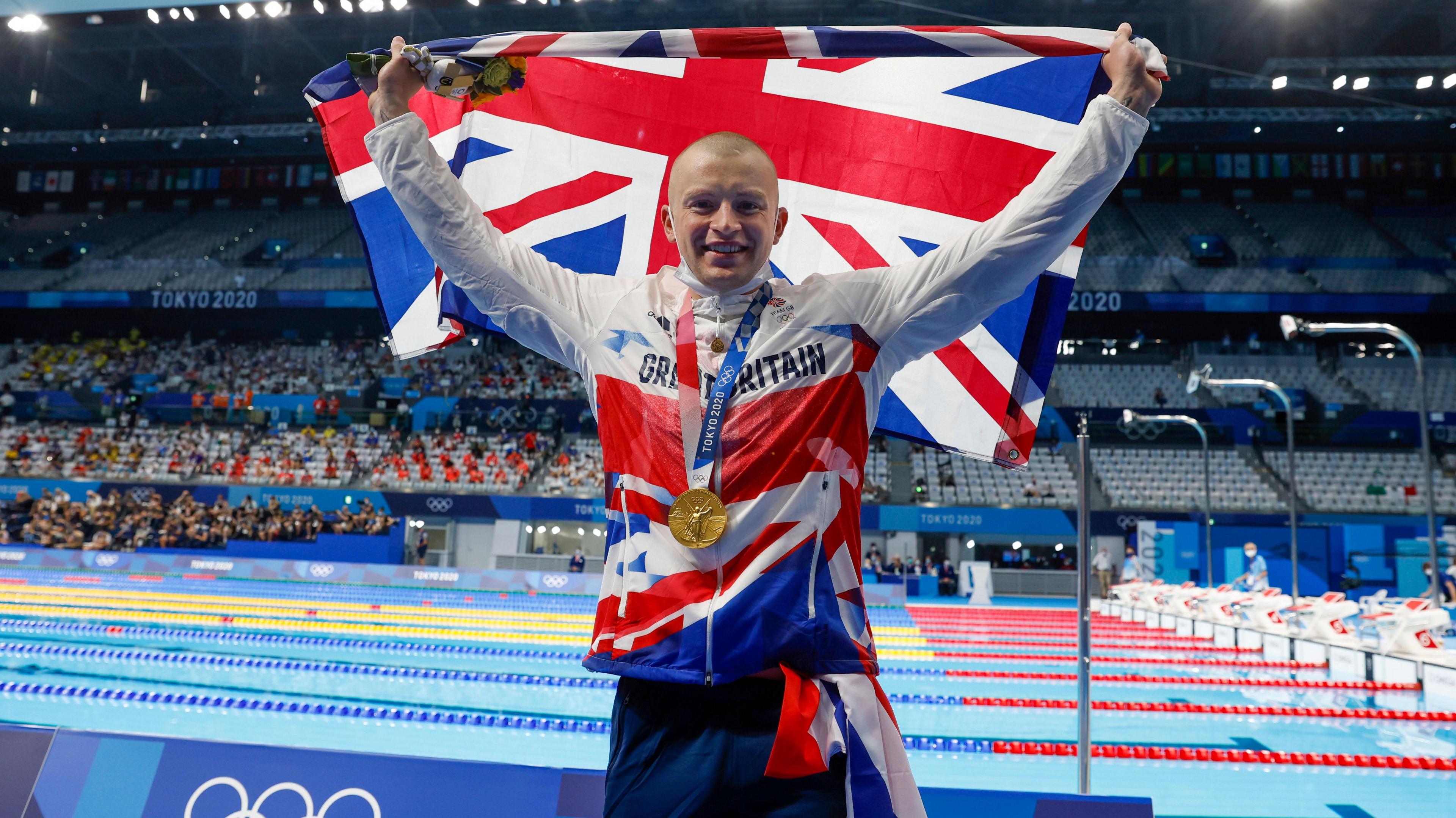 Adam Peaty with a shaved head and wearing a Team GB outfit as well as his gold medal, holds a Union Flag up while he stands next to a swimming pool.