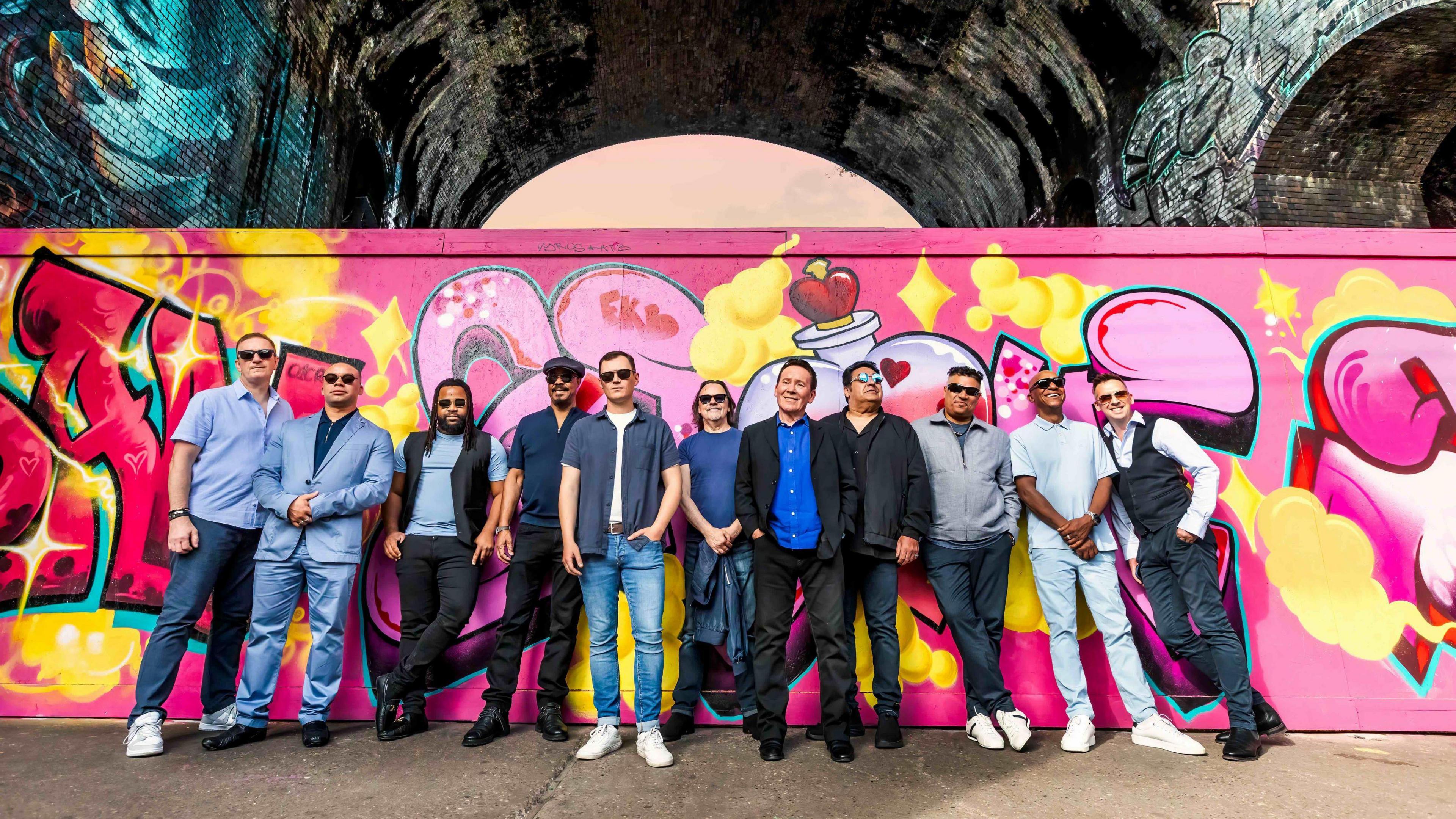 A picture of 11 men standing in a line with pink, yellow and red street art behind them. Some of the men are wearing sunglasses and they are wearing different shades of blue clothes as well as black trousers or jeans.