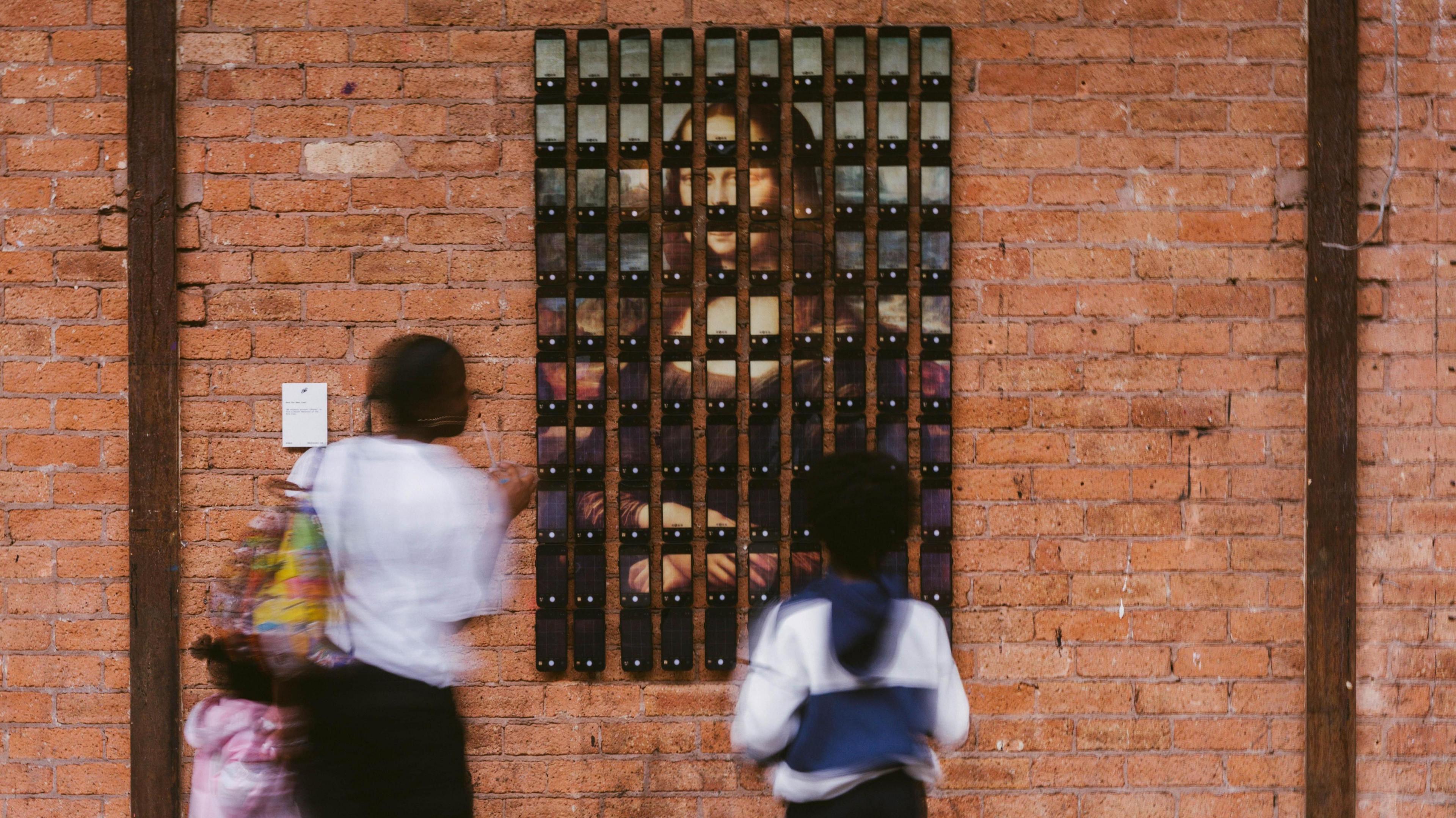 One hundred iPhones are pinned onto a brick wall and each screen shows a snippet of the Mona Lisa portrait. Three people are standing and looking at the display