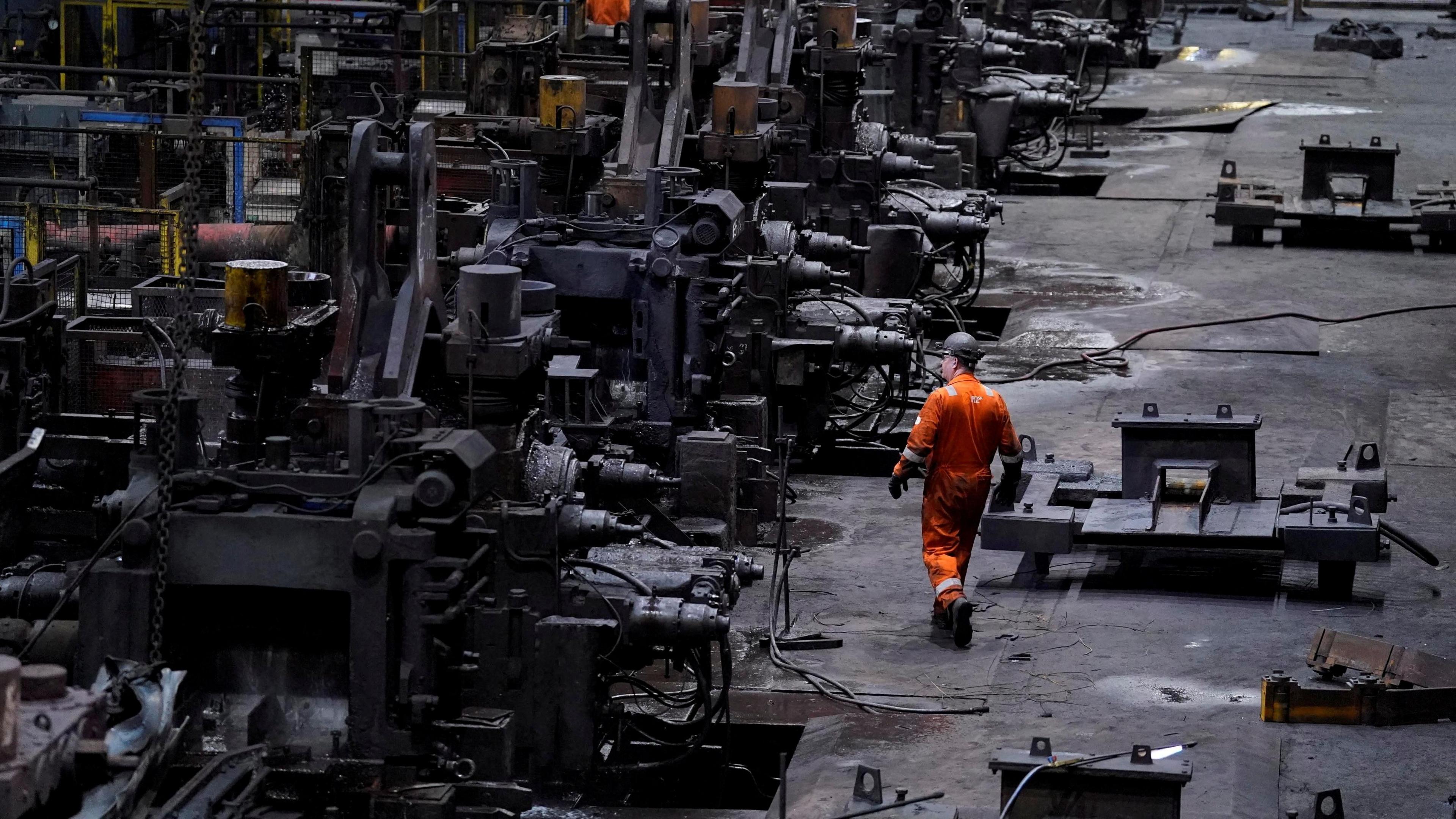 A steel worker dressed in a high-vis orange suit walking through a large steel factory with turbines, large metal, and machinery
