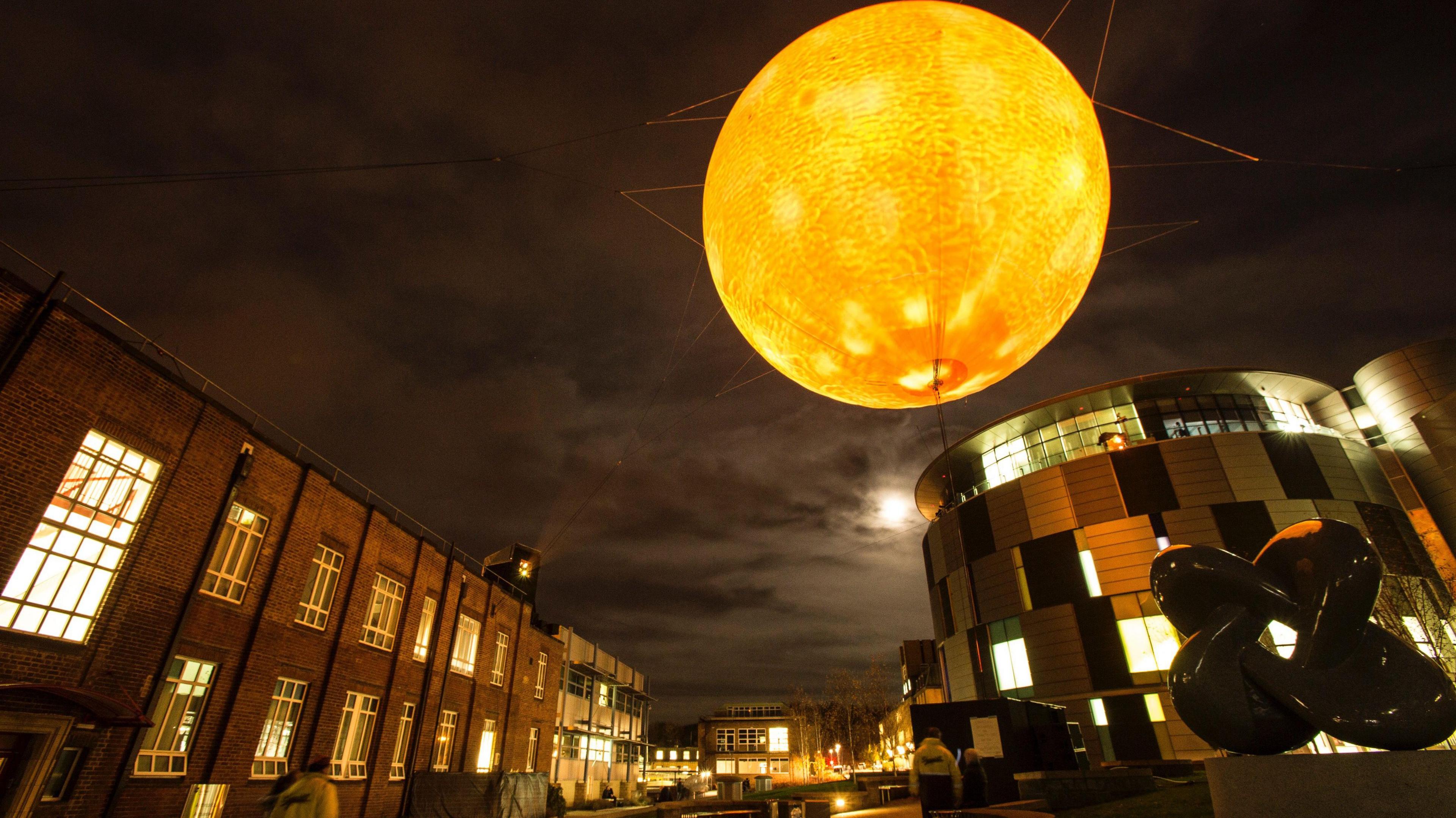Solar Equation is a glowing yellow simulation of the sun, 100 million times smaller. It is suspended above Durham University's Science site, between a modern cylinder-shaped building made of glass and panels and a two-storey brown brick building with white window panes.