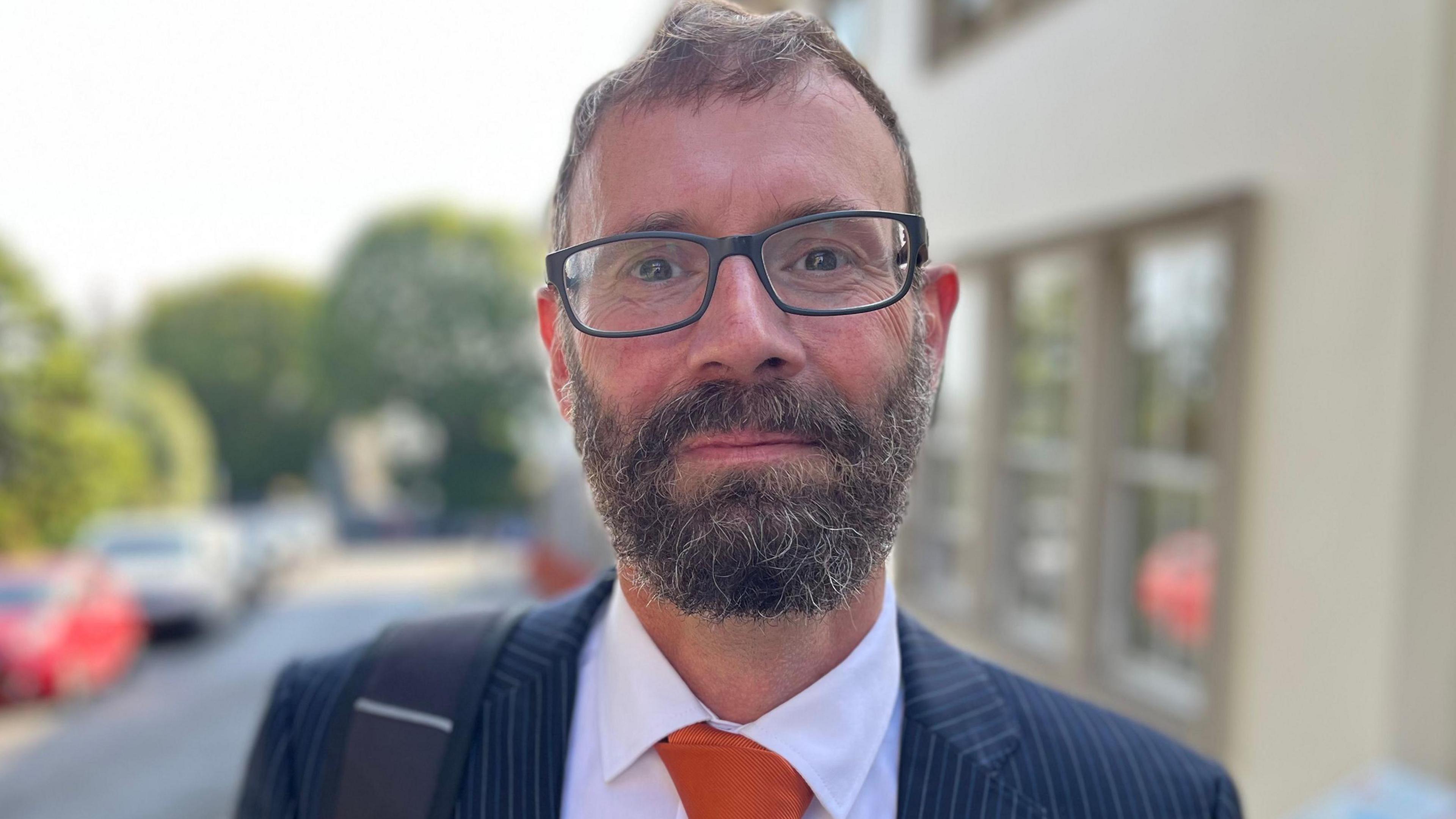 Deputy Aidan Matthews - A man with scruffy short brown hair and a brown and grey beard. He is wearing black spectacles. he has a blue and white pinstripe suit with a white shirt and orange tie. On his right shoulder is a back pack strap. 
