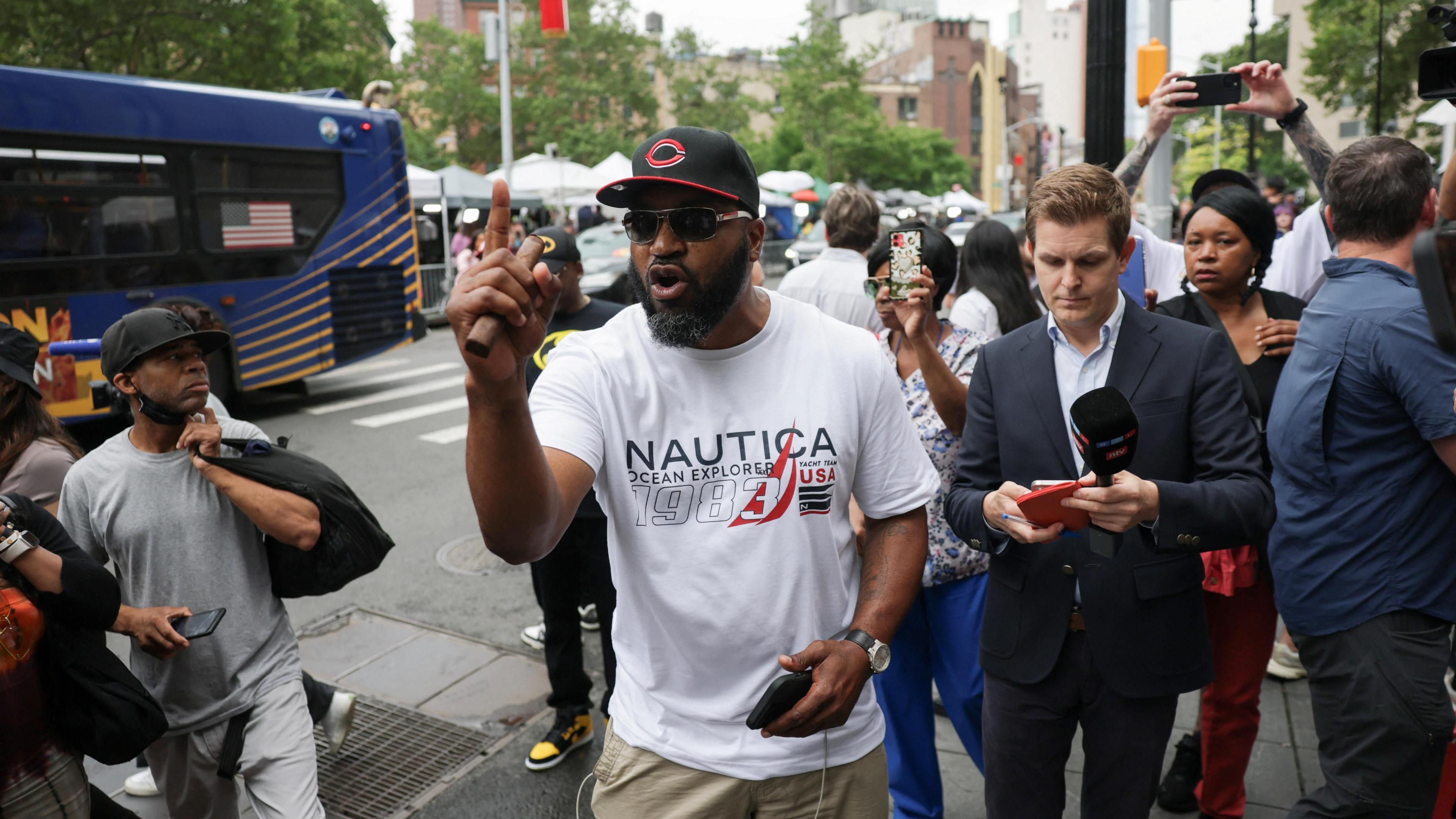 A man wears a white shirt and black hat and holds up a finger in a crowd