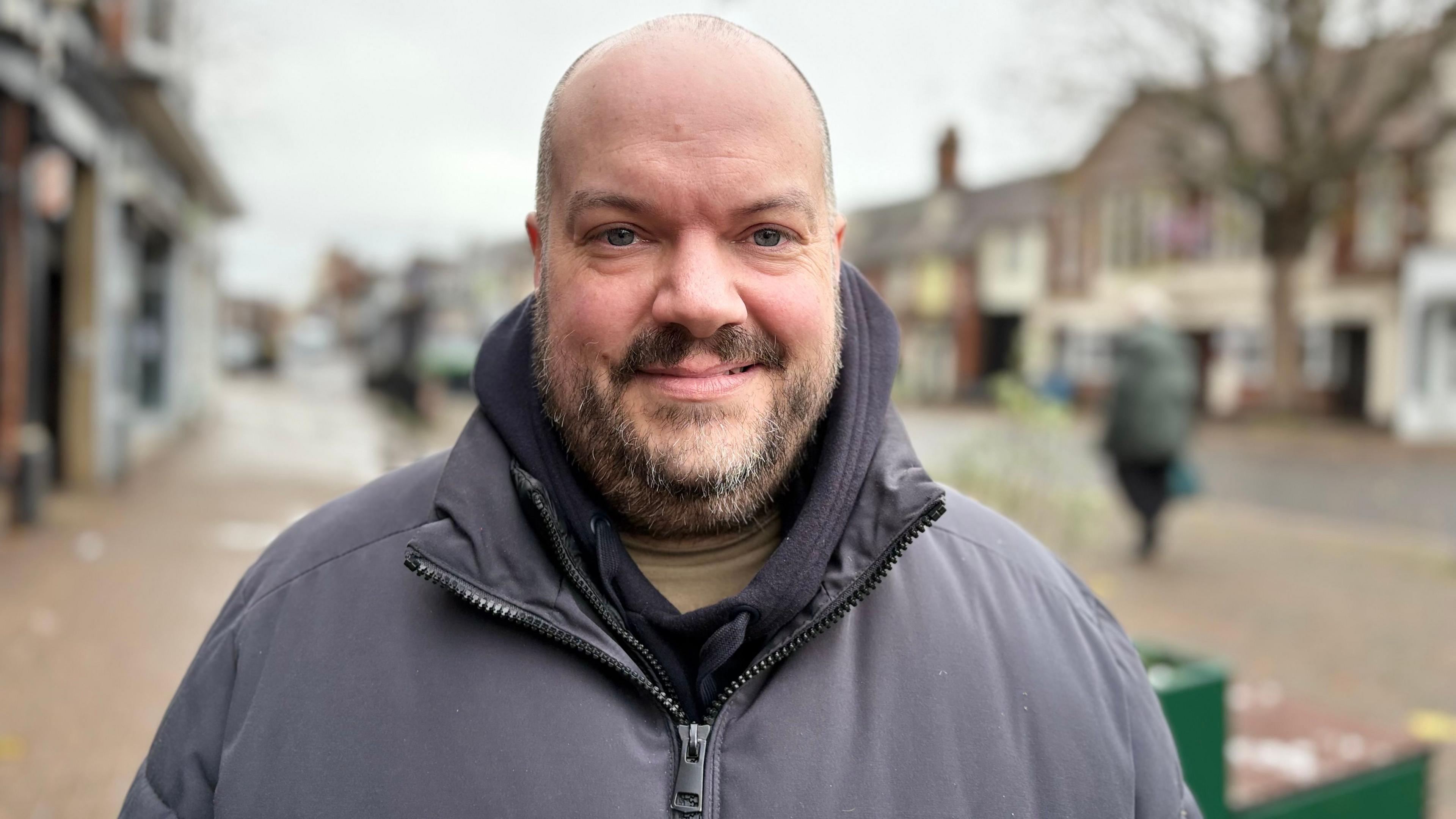 Chris Leaman with very short hair and dark beard smiling at the camera while wearing a grey anorak in Leighton Buzzard High Street with shops in the background and a shoppers in a green coat to the right.