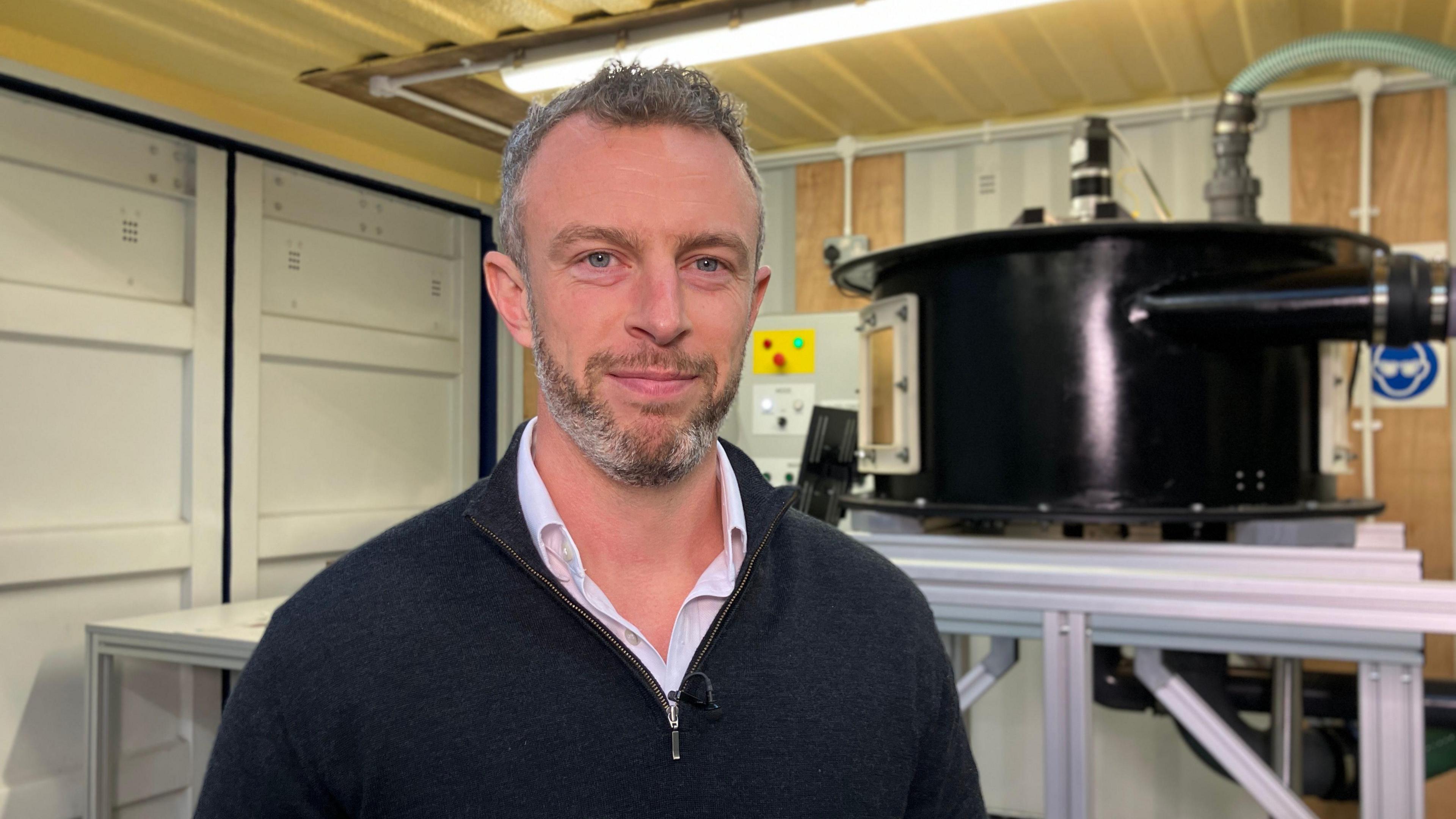 A man is smiling and stood in front of a black system with pipes surrounding it. He is wearing a black fleece over a white shirt.