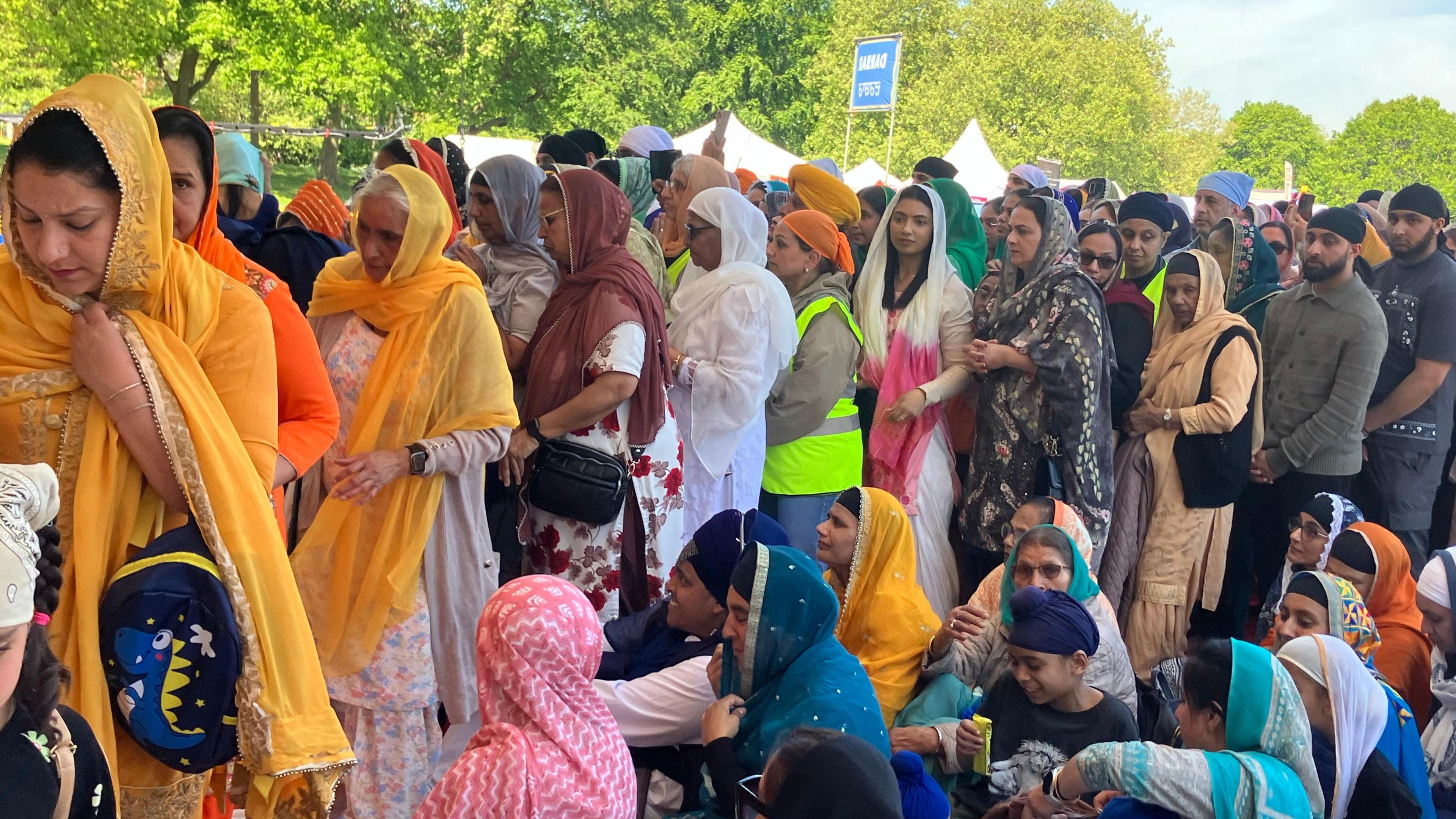 A large group of people dressed in traditional south Asian, that includes women wearing colourful saris, are standing in line. The people in the foreground are sitting on the ground.
