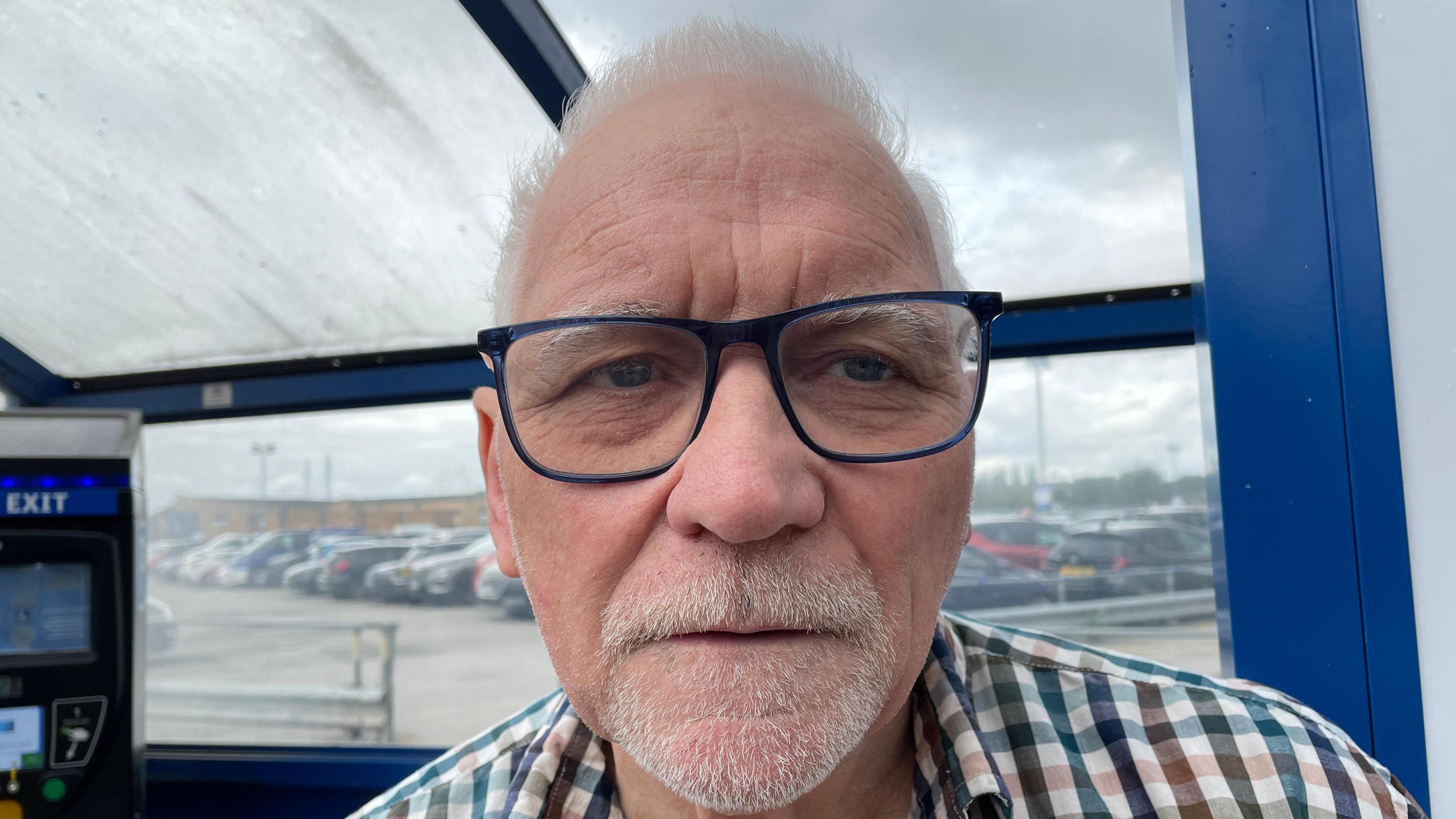 A man is looking at the camera. He has white hair and glasses, and is wearing a checked shirt. Behind him is a parking payment machine.