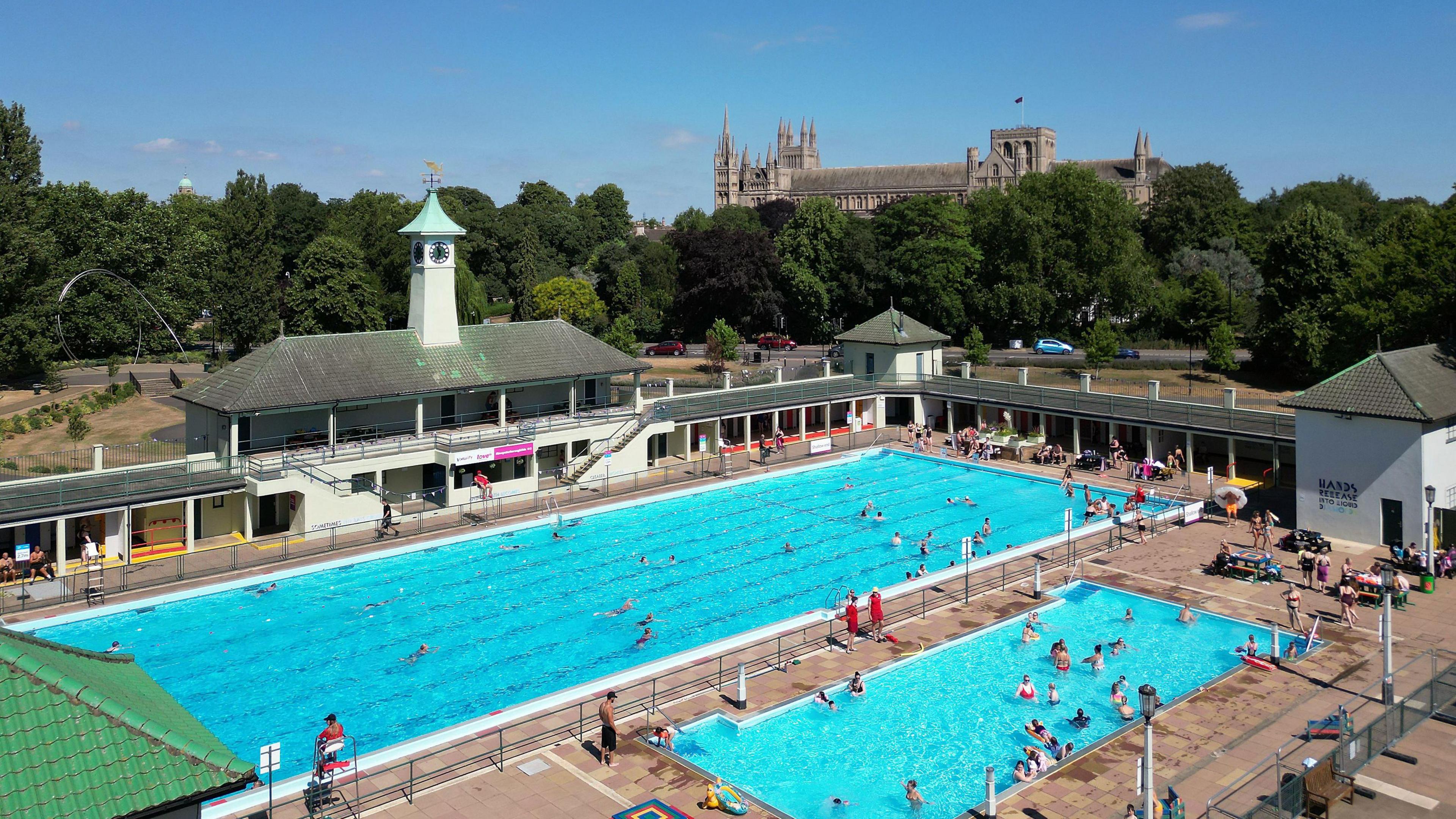 Peterborough Lido, which has two swimming pools - one bigger than the other - with people swimming and lifeguards sitting to the site. The pools are surrounded by white walls with a grey roof and are overlooked by a clock tower with a light blue top. Peterborough's cathedral is obscured by trees.
