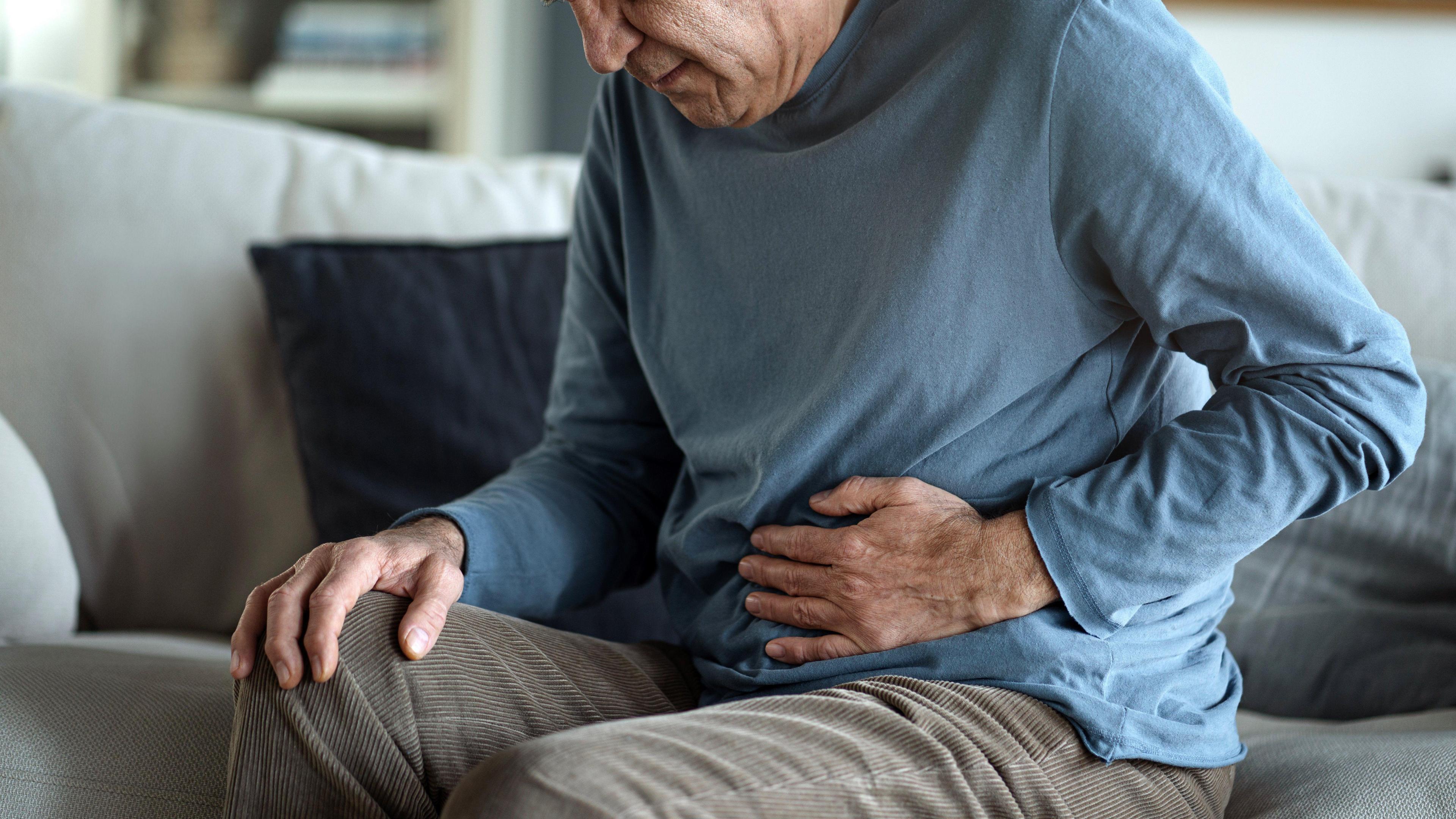 An elderly man is sitting on a sofa with his hand on his stomach. His bent over as if he's in pain.