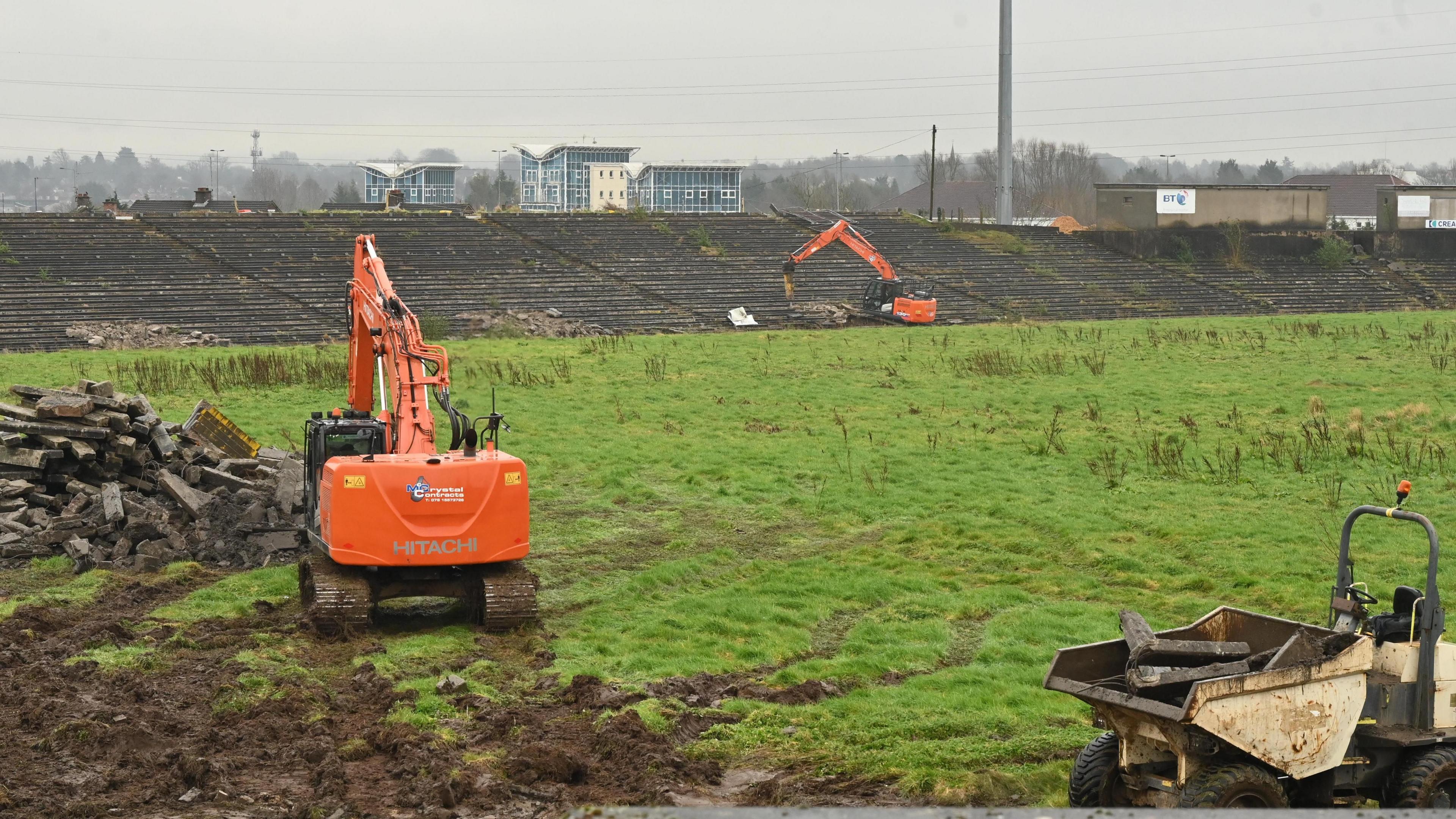 Two orange diggers are clearing the muddy grass of Casement Park under a grey Belfast skyline with bricks on the left and weeds growing out of the pitch. 