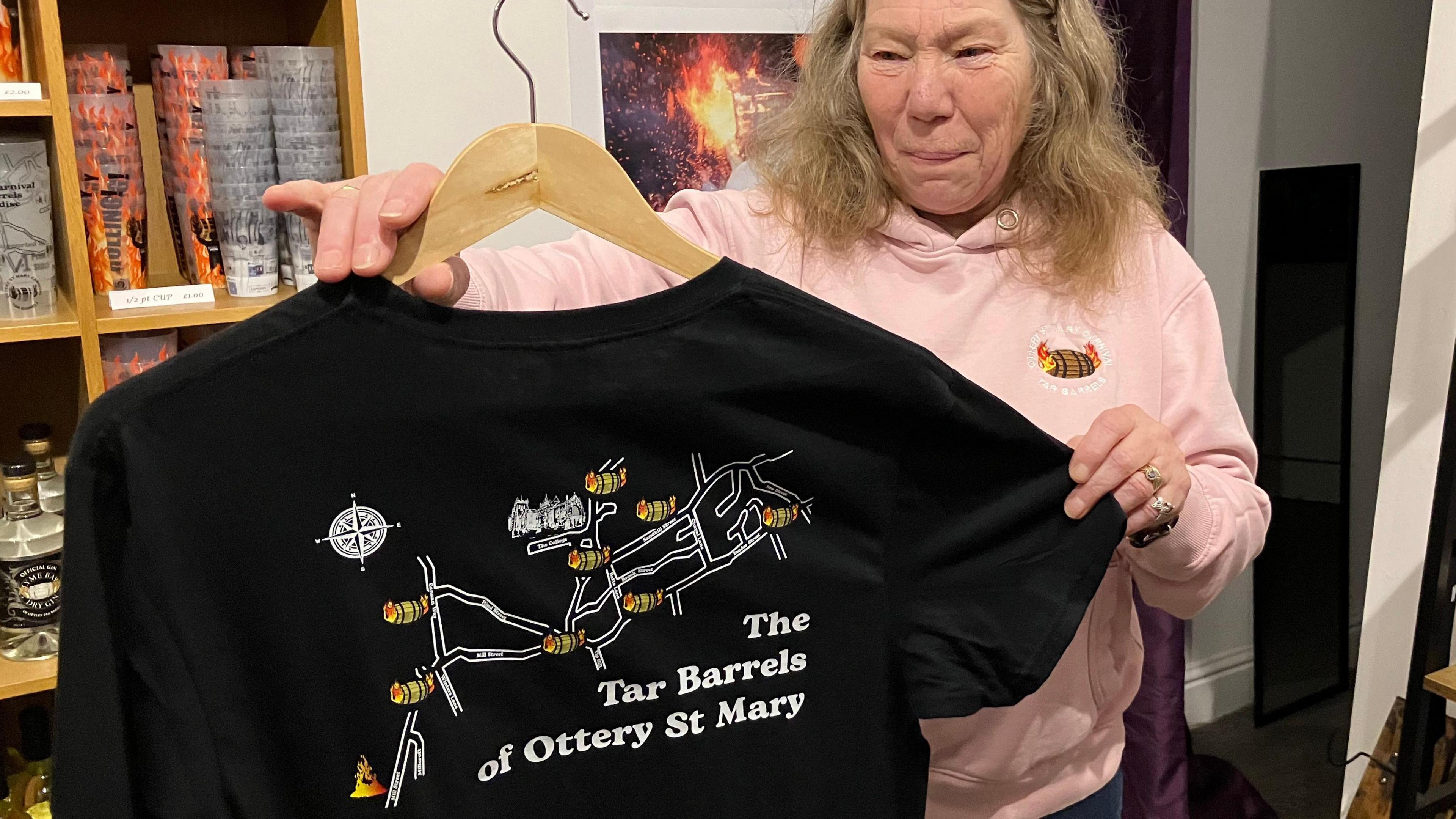 Eva White holding one of the Tar Barrels merchandise t-shirts. She has long brown hair and is wearing a pink hoodie.