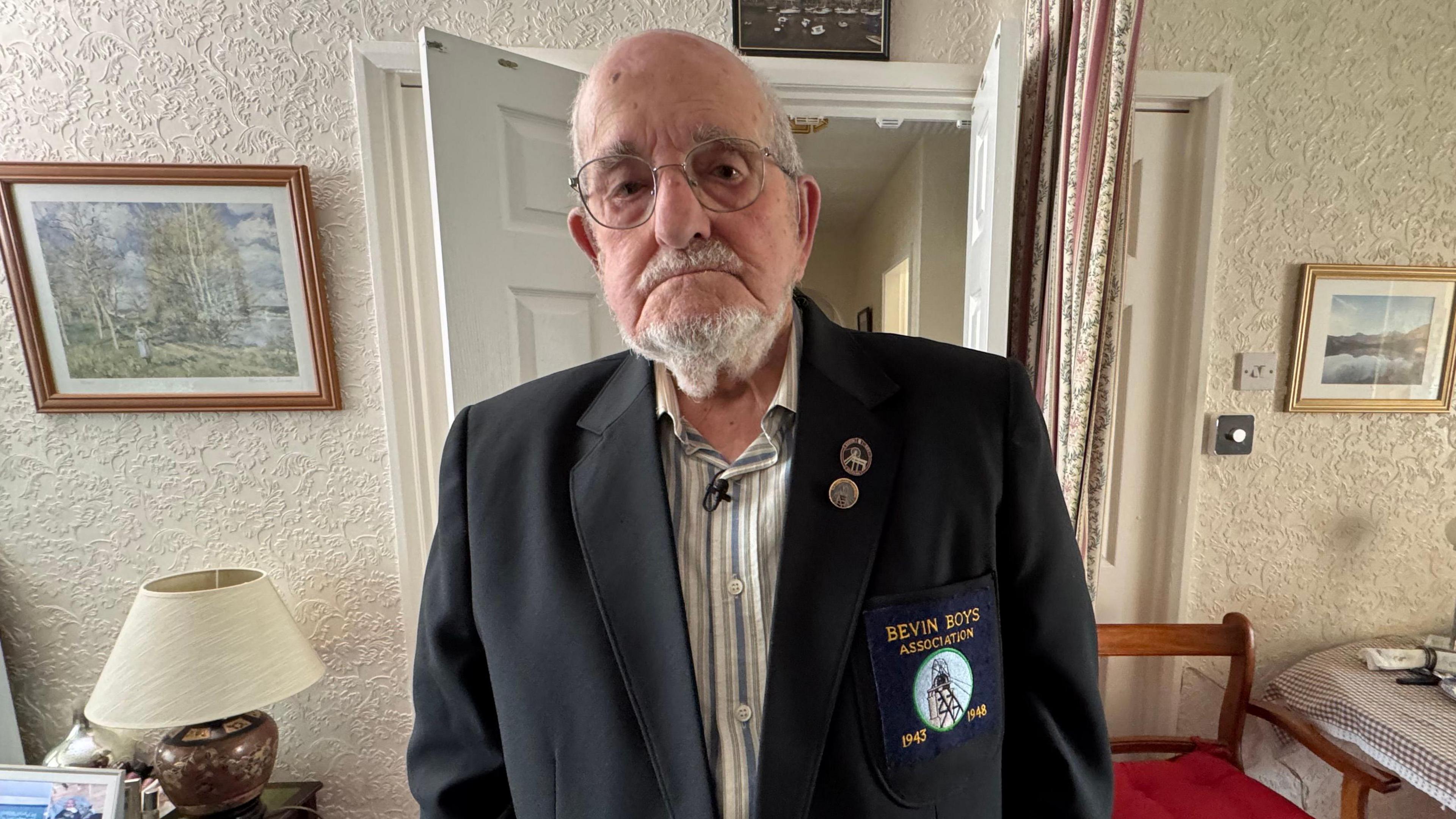 A bald man with a grey beard and moustache, frowning at the camera. He is wearing a white and blue pinstriped shirt and a navy blazer with a "Bevin Boys Association" logo. Behind him is a living room with a table, chair and lamp and pictures hanging on the walls.