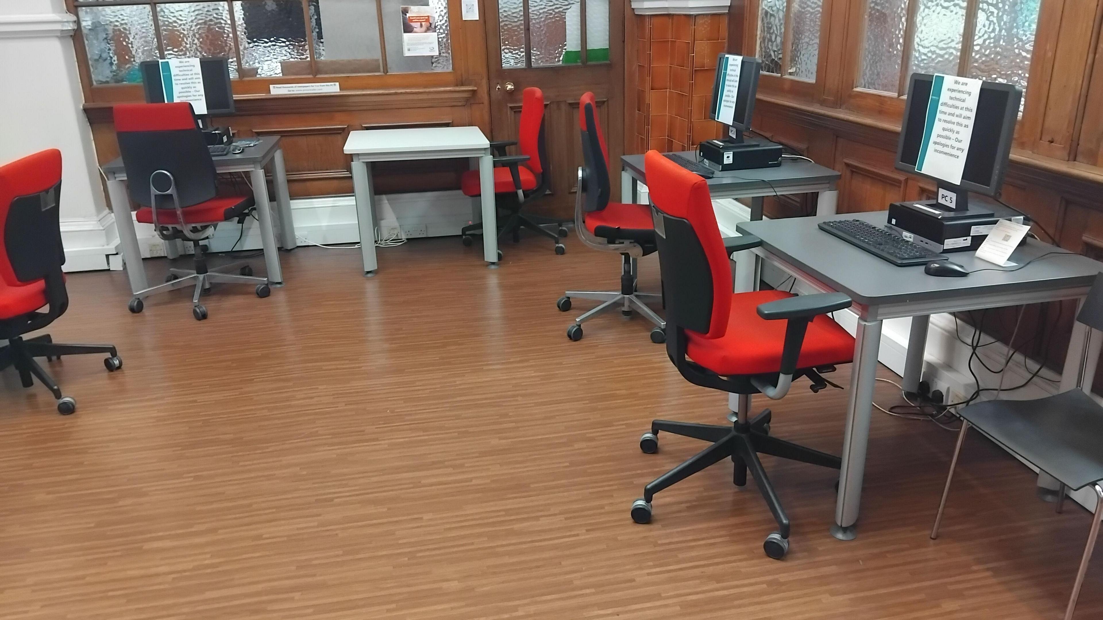 A room in an Edinburgh library in which all computers are turned off, with notices on the screen detailing they cannot be used due to a 'technical fault'. The office chairs pulled up the at the desks on the dark wooden floor are red, with a black back and frame.