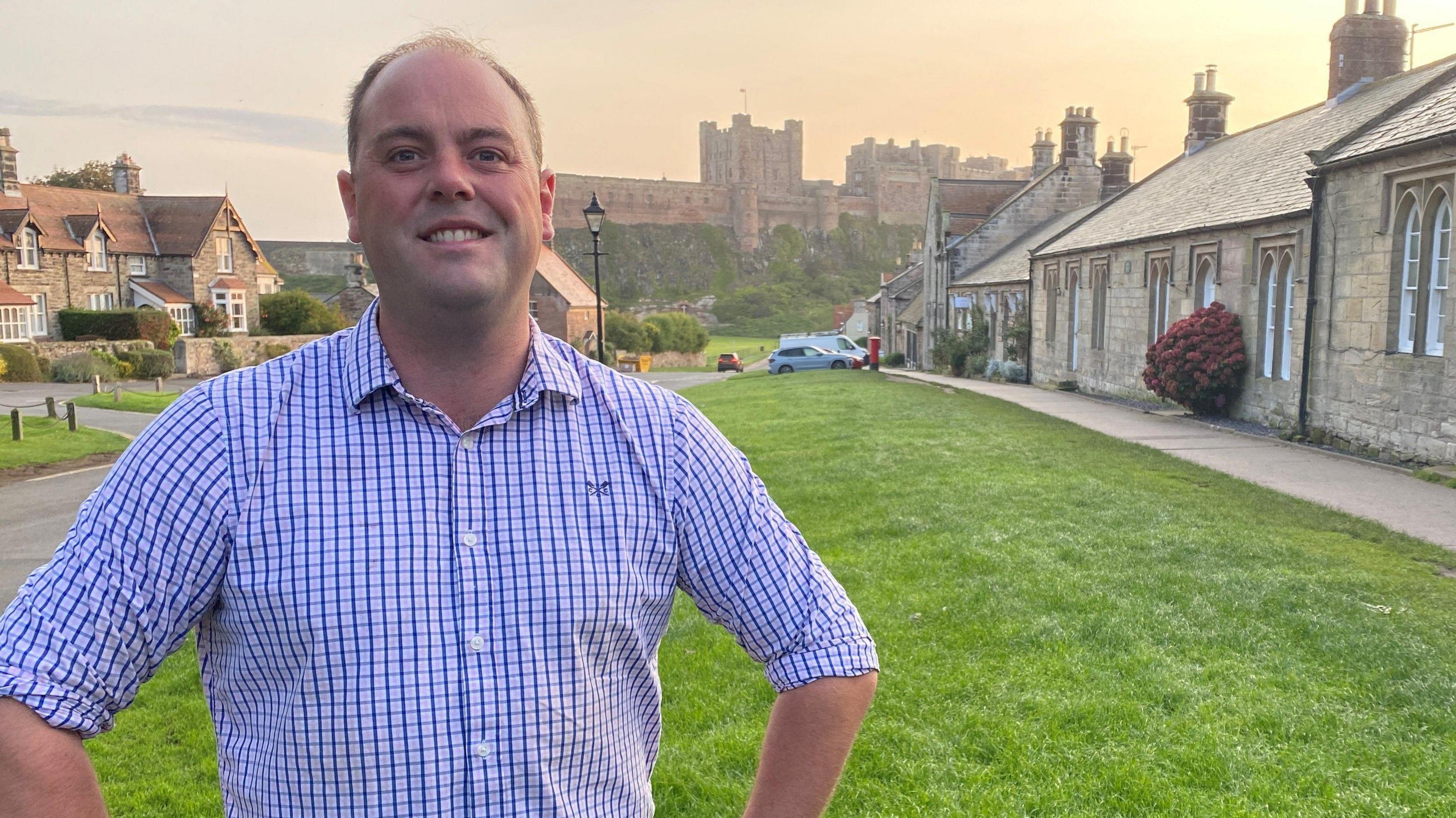 Guy Renner-Thompson is standing in front of Bamburgh Castle. He is a man in his 40s, with a receding hairline and wearing a blue checked shirt.