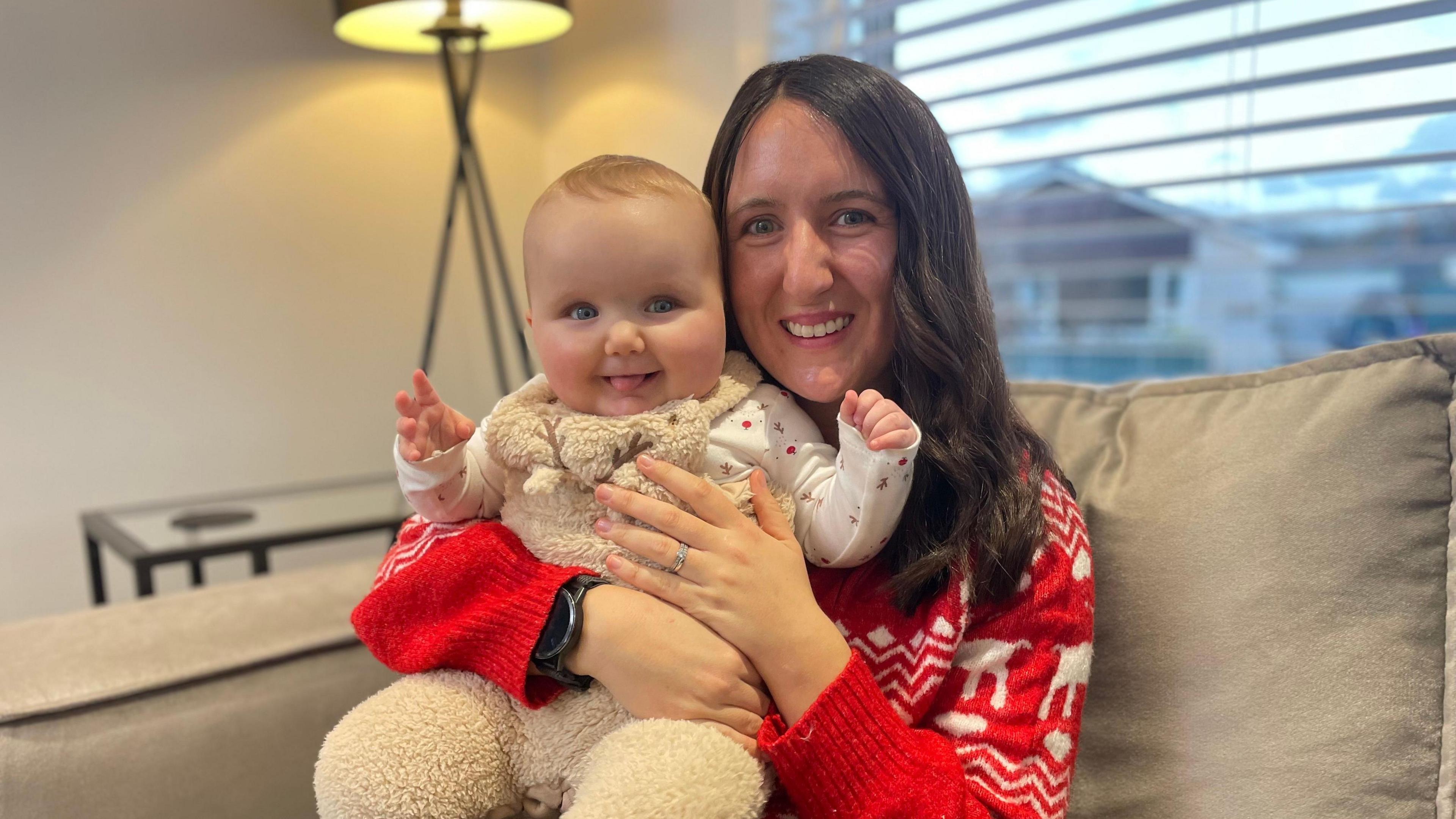 Kirsty Mathieson holding her baby daughter Gia. They are both looking straight at the camera. Kirsty has long dark hair and is wearing a red and white Christmas jumper. Her daughter is wearing a furry reindeer Babygro. 