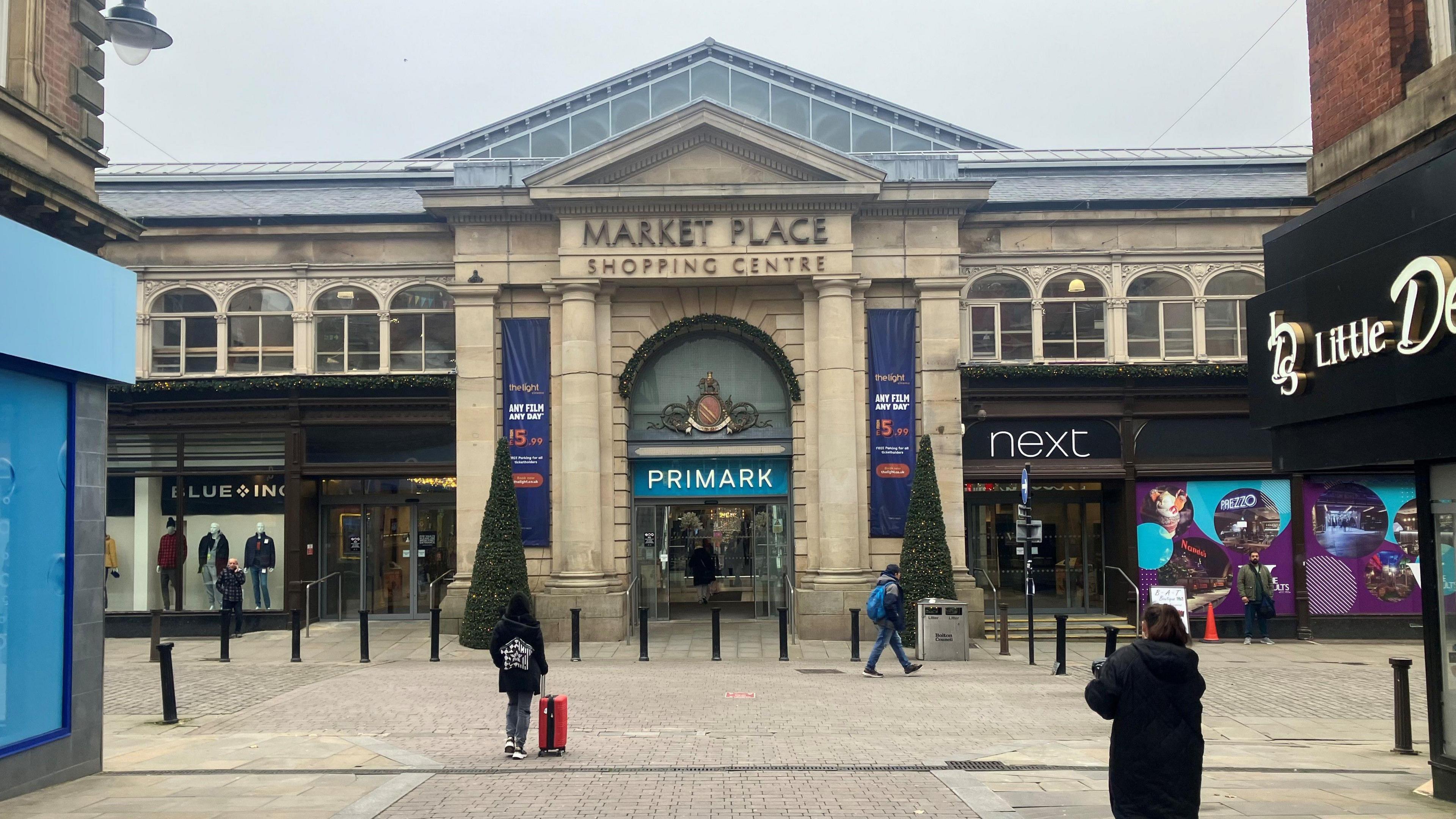 Bolton's Market Place shopping centre, with shoppers on the street. Prmark is at the heart of it - next to Next and two christmas trees flank the entrance. 