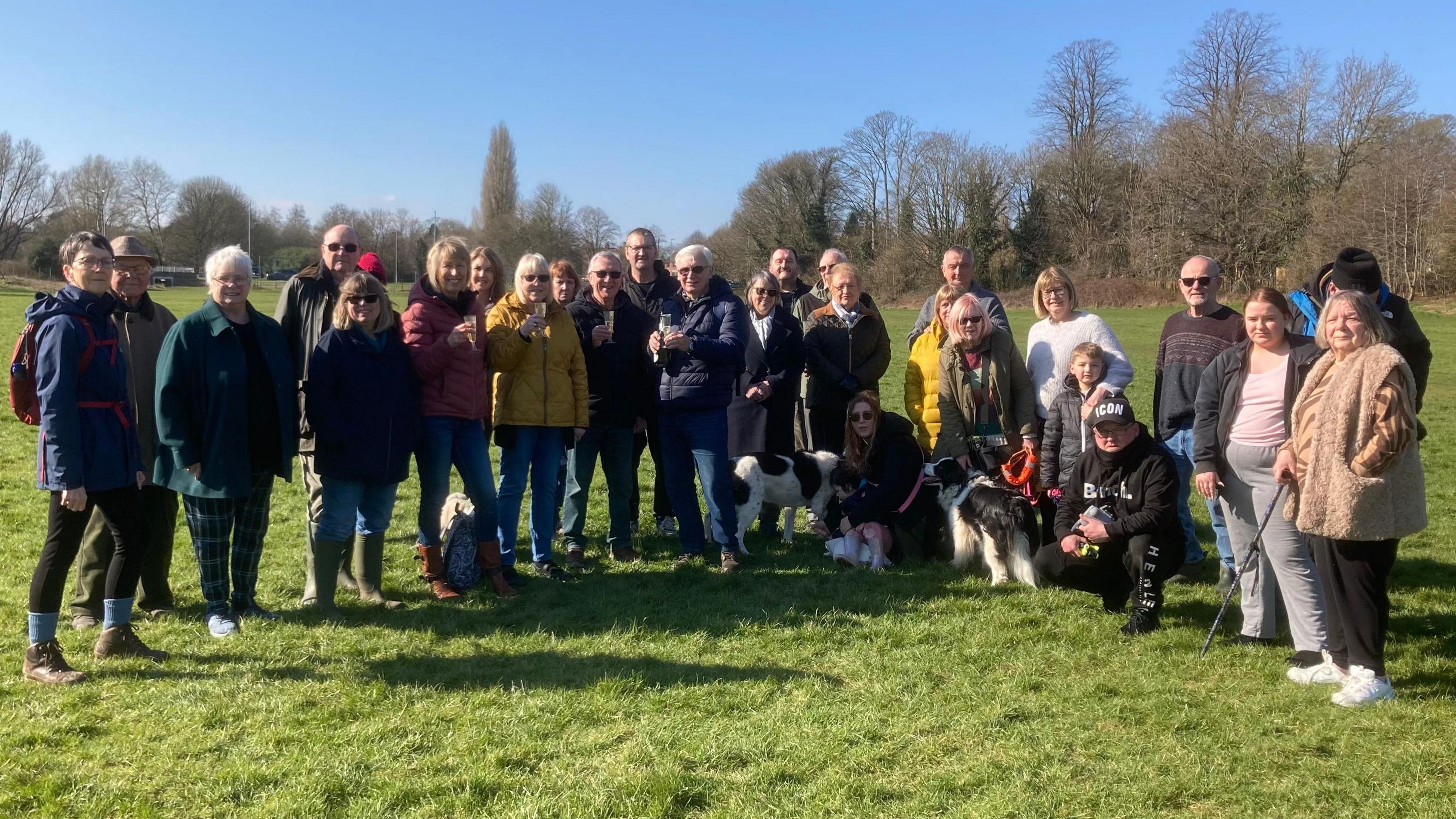 About 30 people standing in a semicircle of various ages. Many are wearing anoraks. There are four dogs within the group.  They are standing in a grass field with trees behind them.