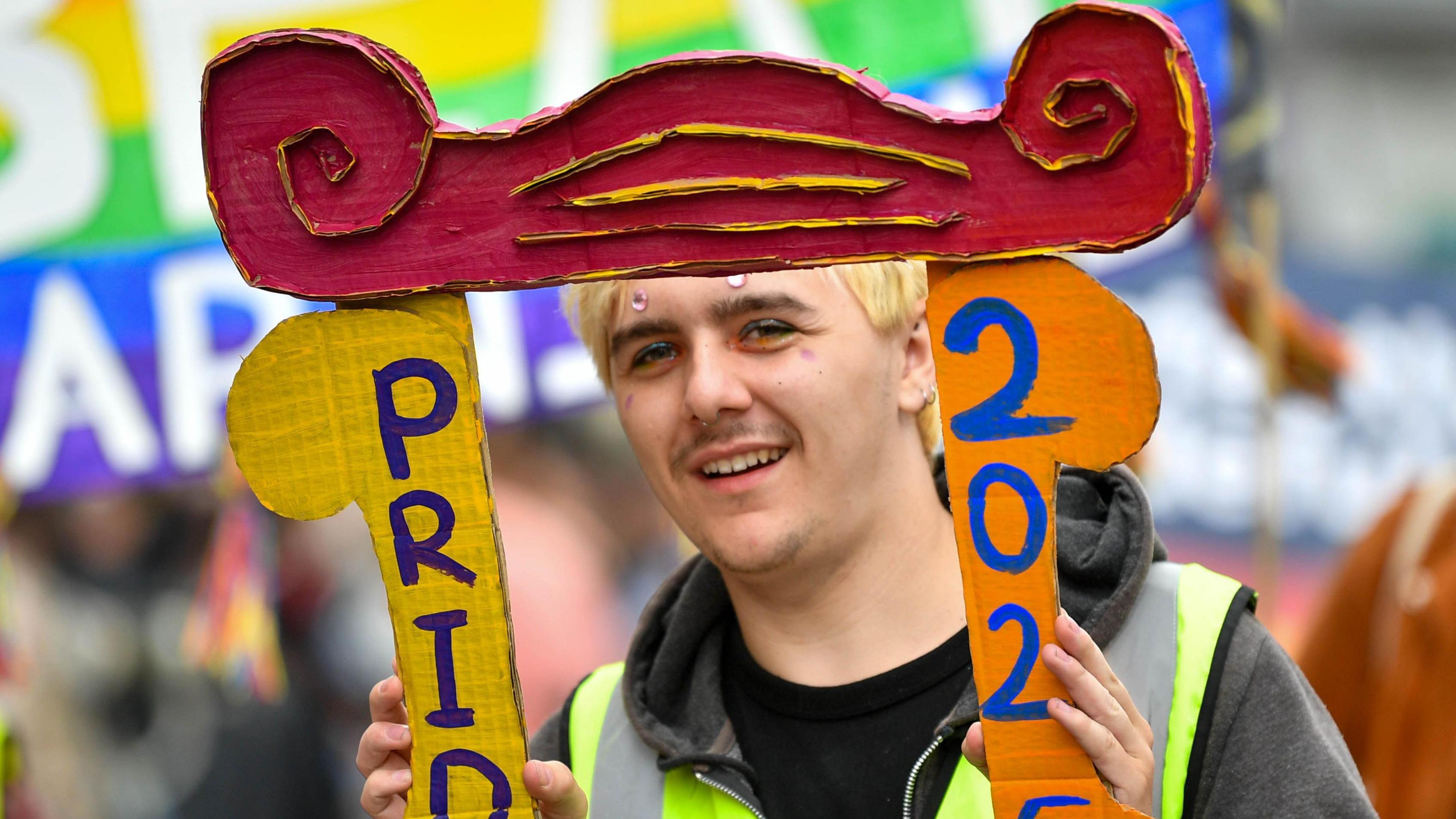 Someone is a parade is looking through a large frame that says Pride 2025.