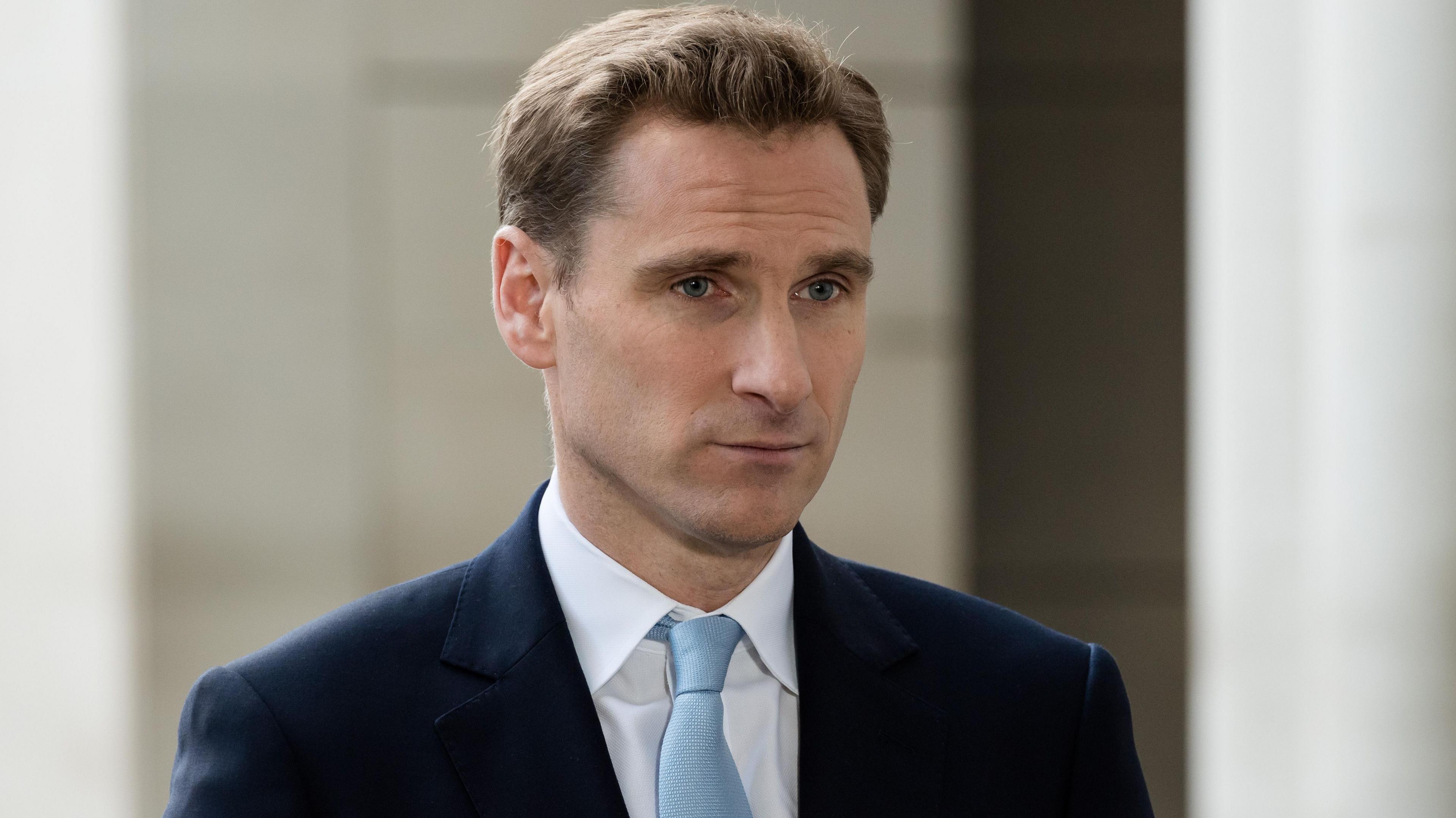 Shadow home secretary Chris Philp, pictured in a dark blue suit and light blue tie. He has a side parting in mousy brown hair and is looking directly forward.