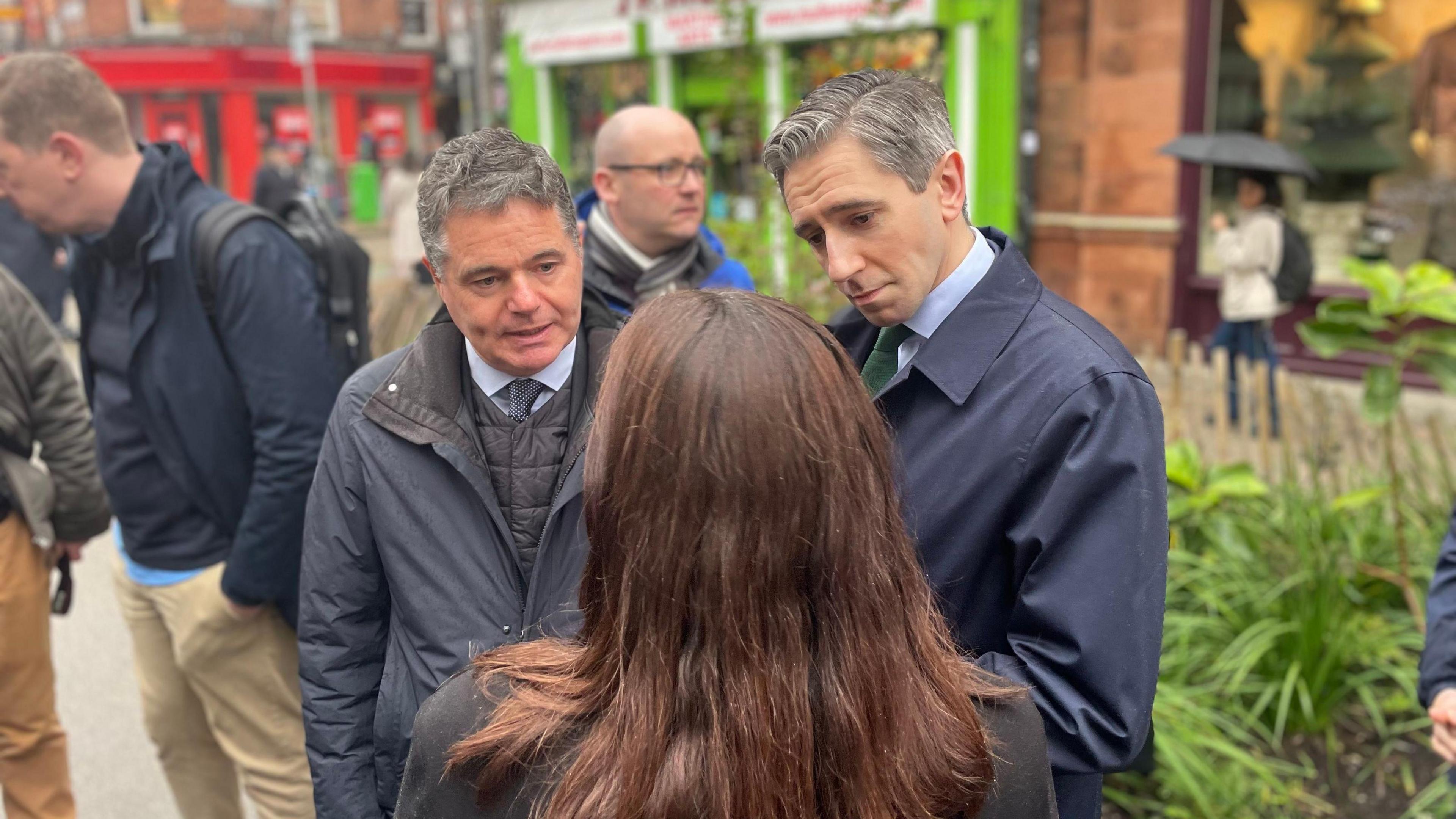 Paschal Donohoe standing beside Taoiseach Simon Harris. Both men are wearing blue shirts and navy jackets. A woman with brown hair is standing in front of them, facing them. The back of her head is visible in the photo. They are standing on Capel Street in Dublin, which is visible in the background of the image. 