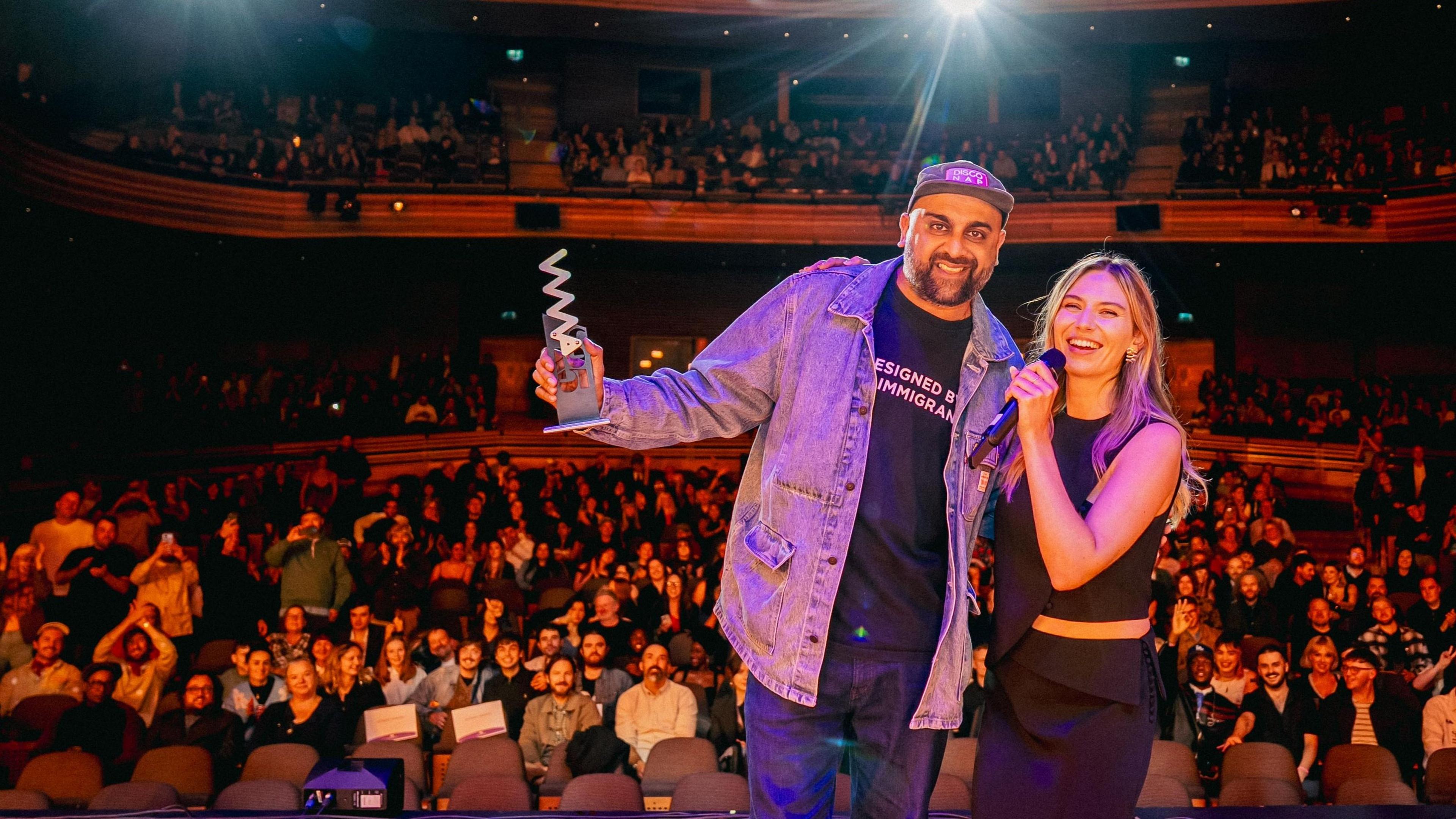 Aly Jamal or Don Leisure pictured on stage at the Wales Millennium Centre, with the audience applauding behind, receiving his award from an unnamed female presenter.