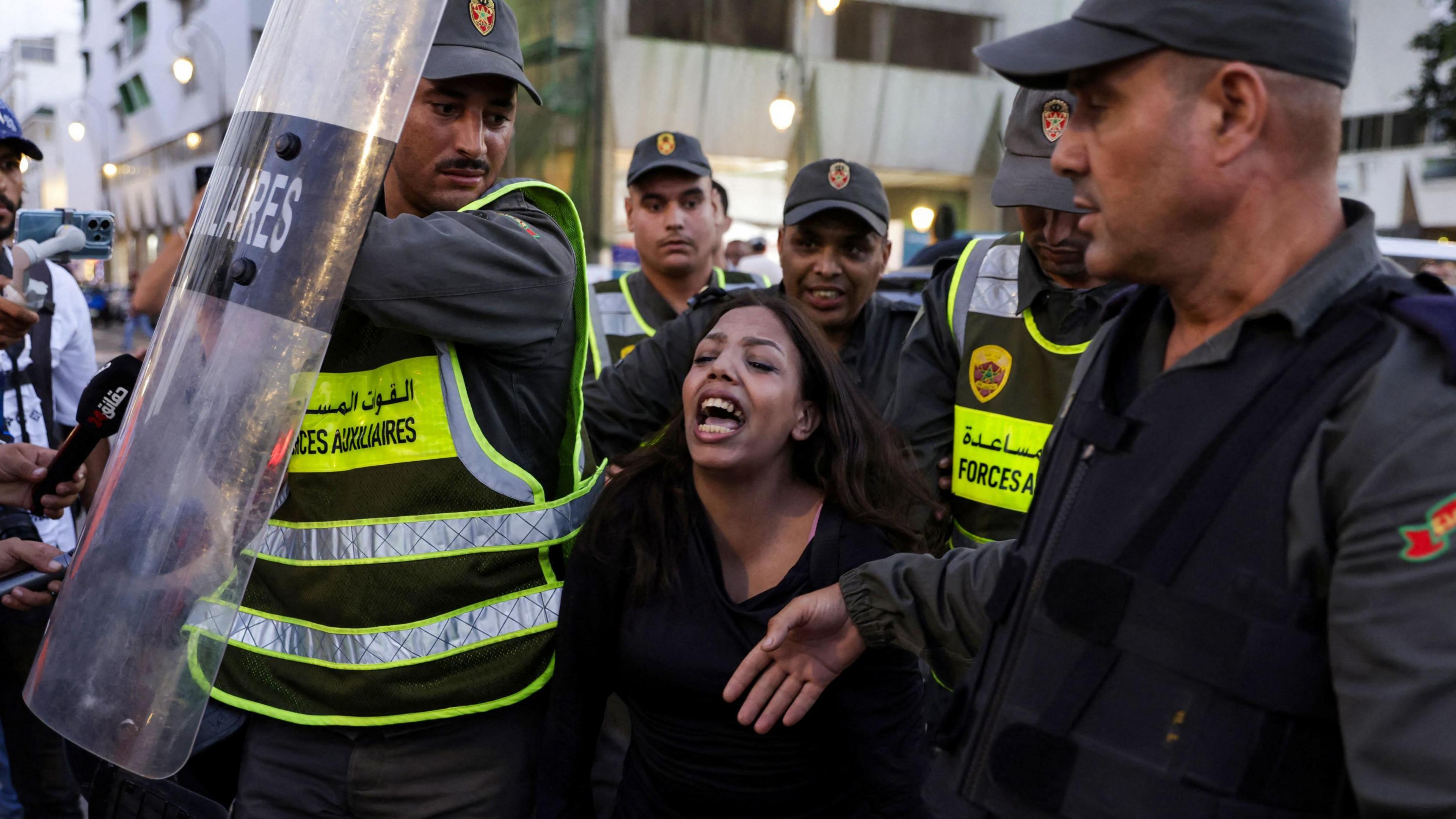 A woman is arrested by police officers, who are surrounding her. One is holding up a plastic riot shield
