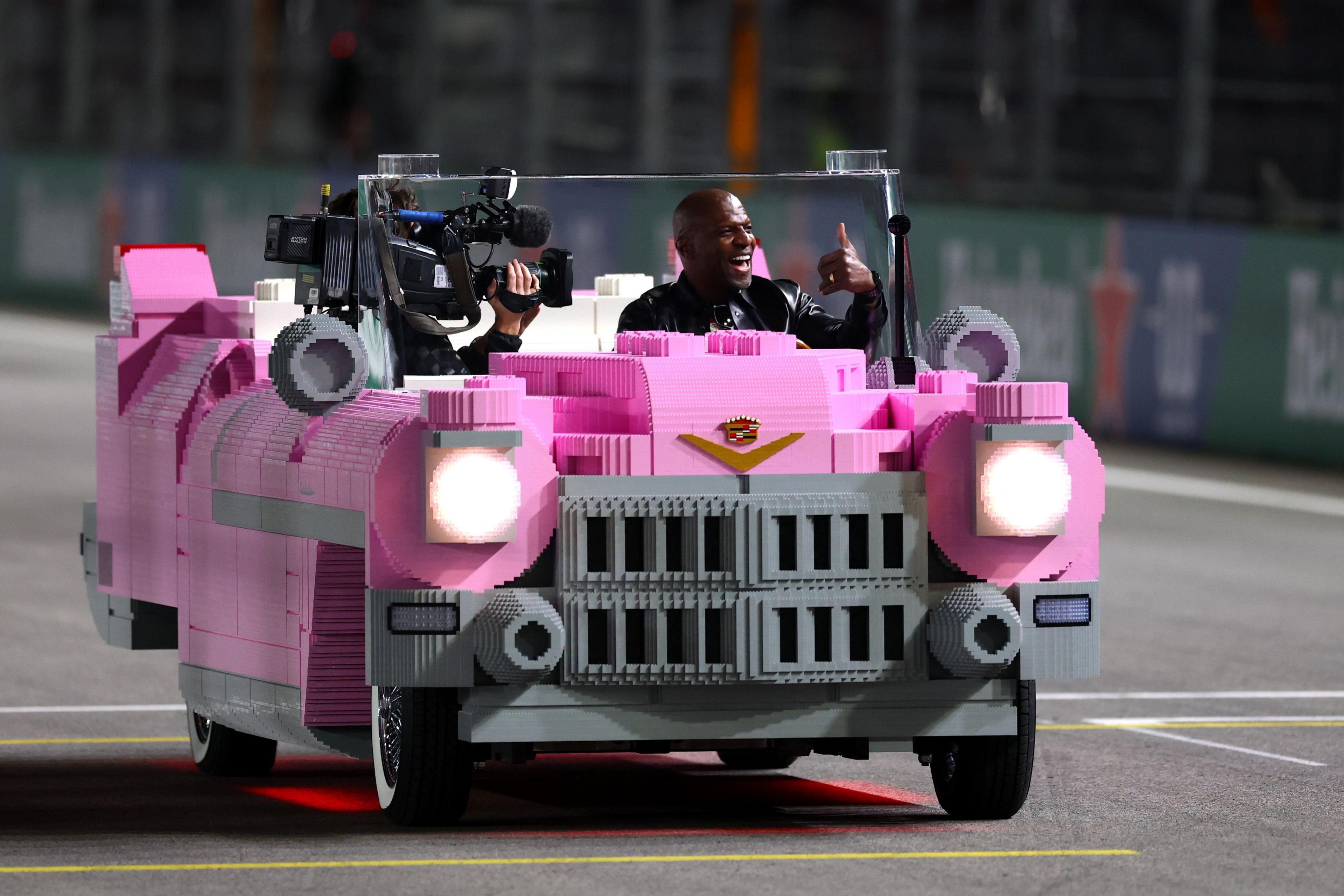 A bright pink Lego-style Cadillac is driving on a racetrack, equipped with cameras mounted on the front. The car has detailed blocky construction, headlights and a Cadillac emblem, with barriers and advertisements visible in the background.
