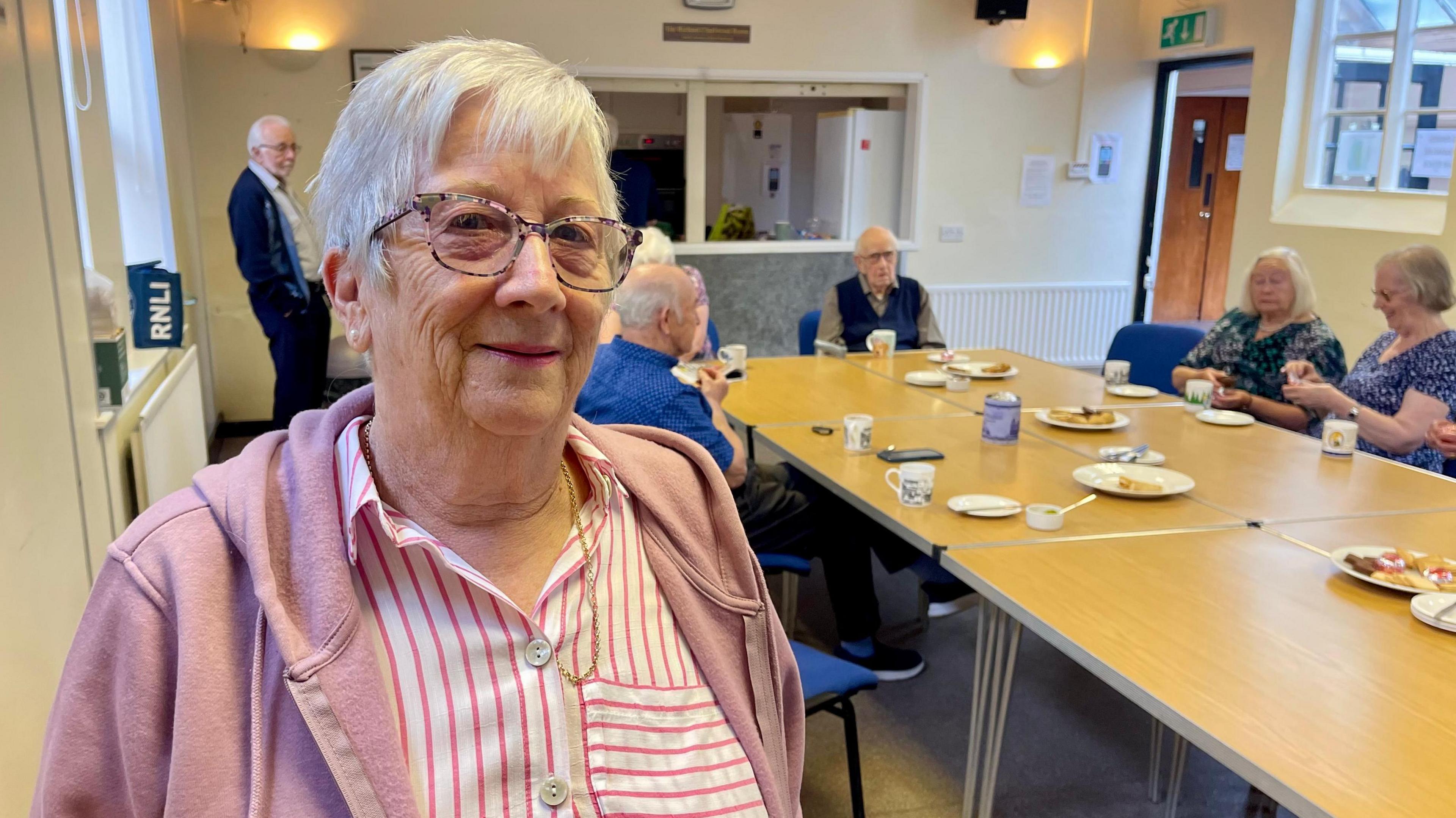 A woman with white hair is wearing glasses, a pink hoodie and a white/pink striped shirt. She is standing inside a village hall with five people sitting around a wooden table behind her and one man standing in the corner. There is a large hatch that leads to a kitchen area. There are cups of tea and plates of biscuits on the table.