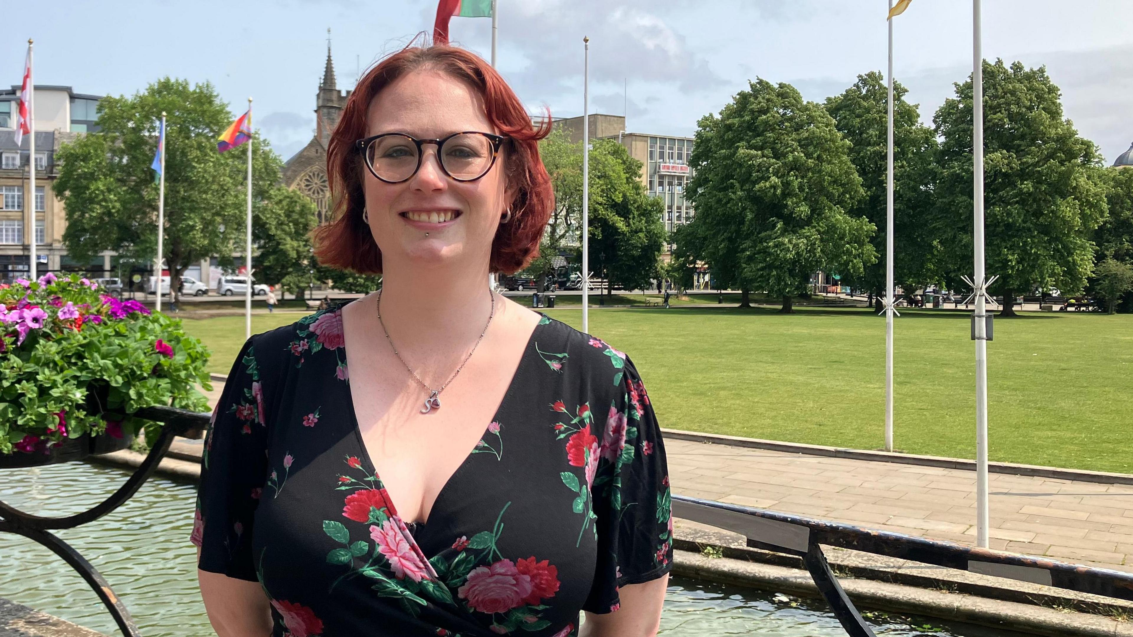 Sarah stands outside City Hall, smiling at the camera. She has short red hair and wears a black floral dress and round glasses. Behind her is a railing with a flower basket on it, part of a large water feature, several flagpoles and the green expanse of College Green. She is smiling at the camera and has her hands clasped behind her back.