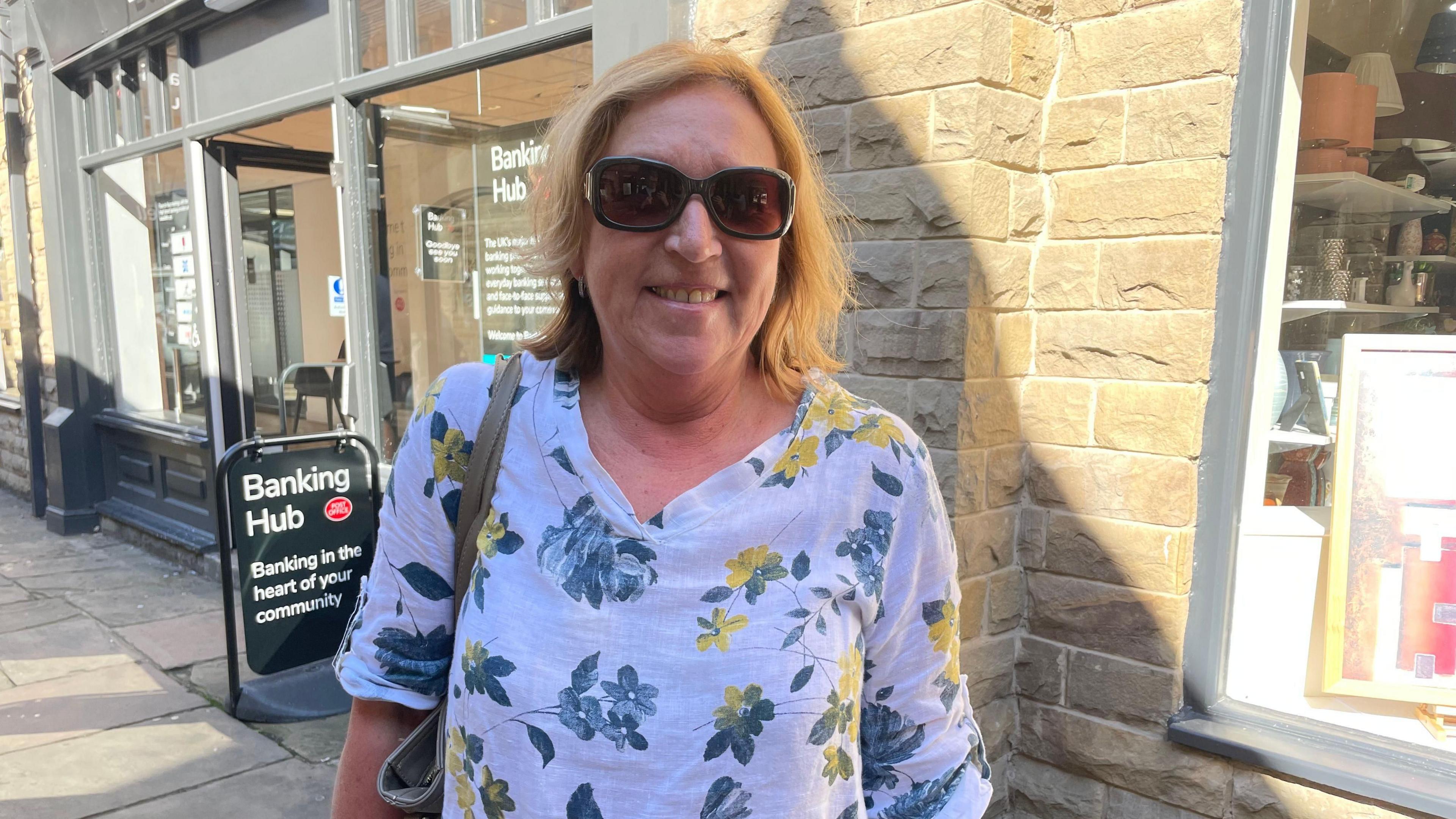 A woman wearing a white floral print top and sunglasses stands in front of a sign for a banking hub