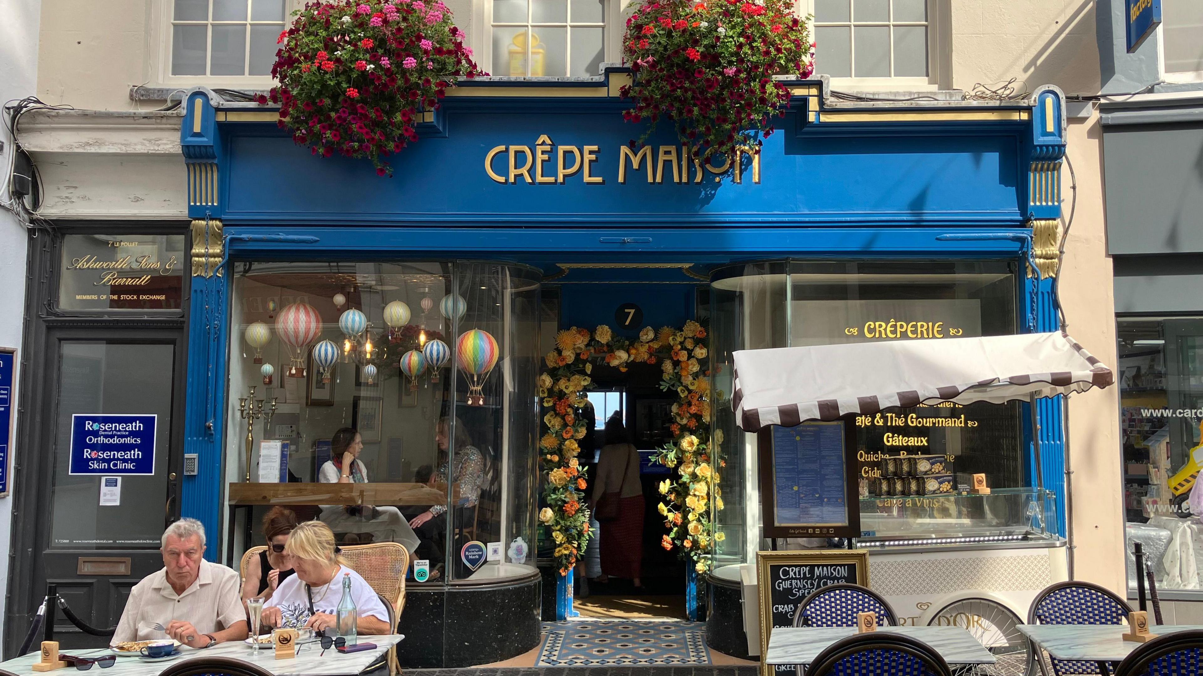 Another street scene, featuring French-inspired creperie, 'Crepe Maison' - in a deep blue colour, with gold lettering. Pink flower baskets hang from the windows, as customers enjoy crepes and bakery goods on the tables outside. 