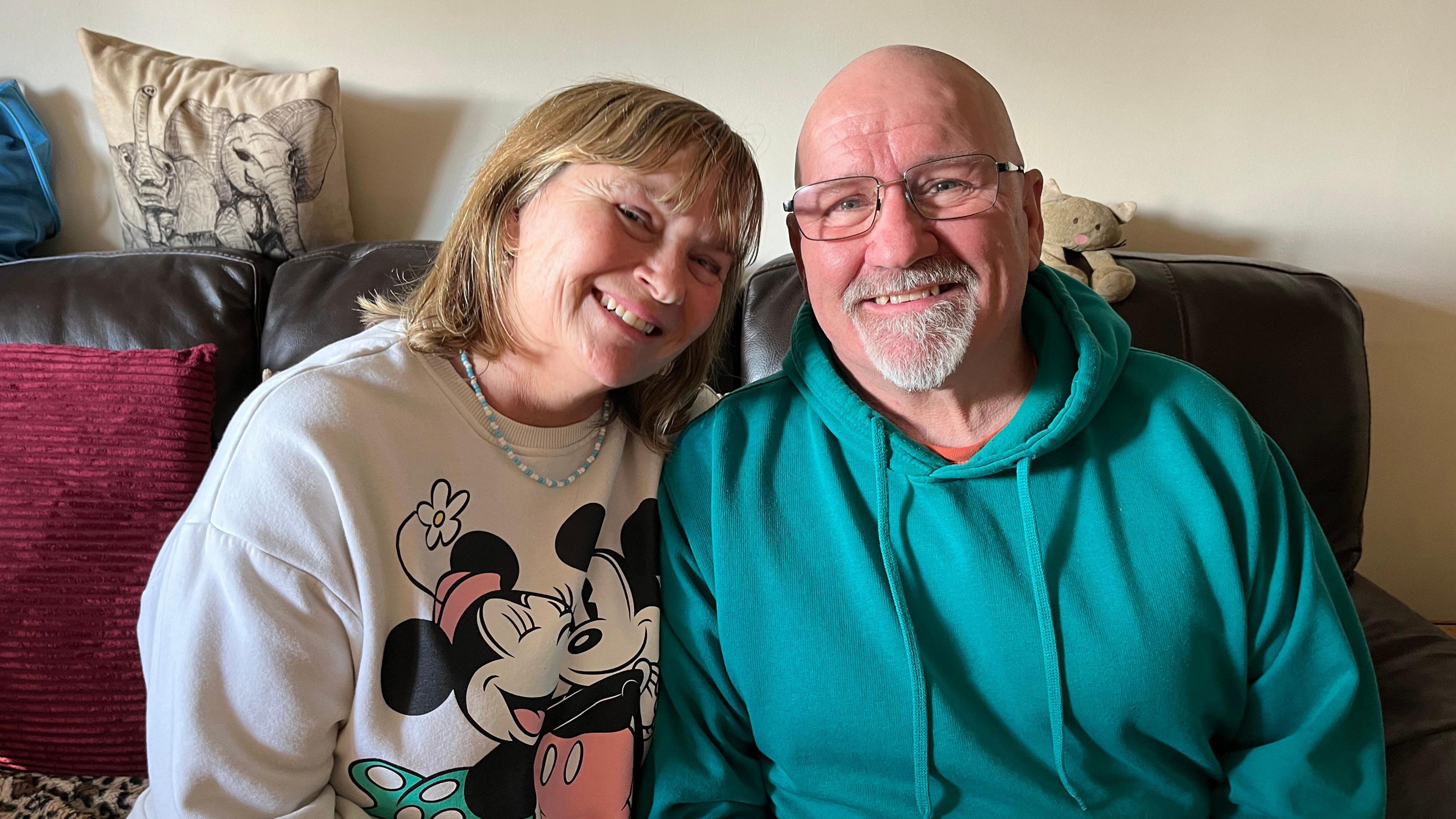 Tracey and Mike Lane are sitting on a brown leather sofa. Tracey has dark blonde hair. She is wearing a Mickey Mouse jumper and is smiling. Mike is wearing a blue hoodie.