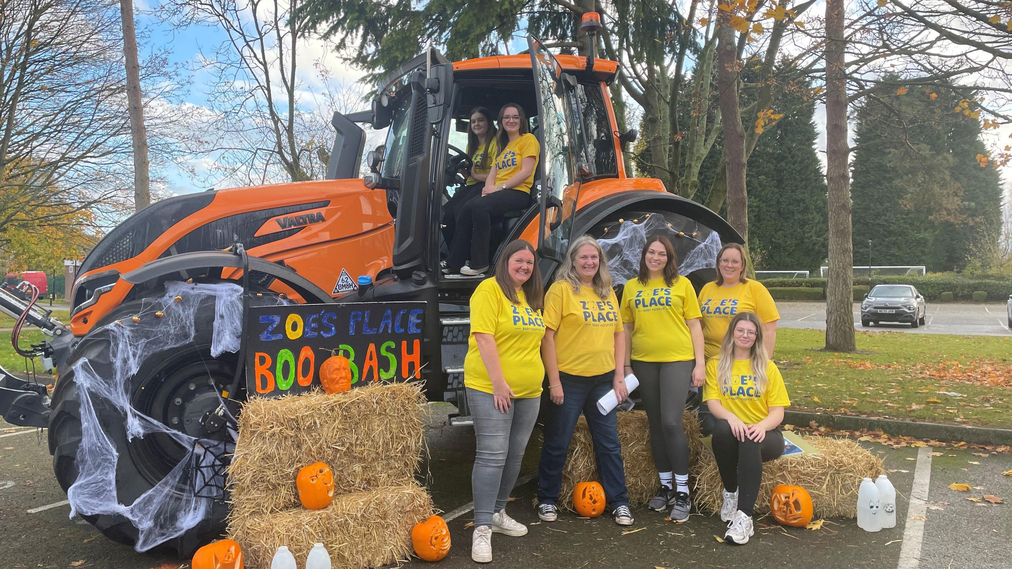 Five women wearing yellow t-shirts with "Zoe's Place' written on. They are standing or sitting on haystacks and there is also an orange tractor with net shaped like cobwebs on it and the word "Zoe's Place Boo Bash" in blue, orange and green writing.