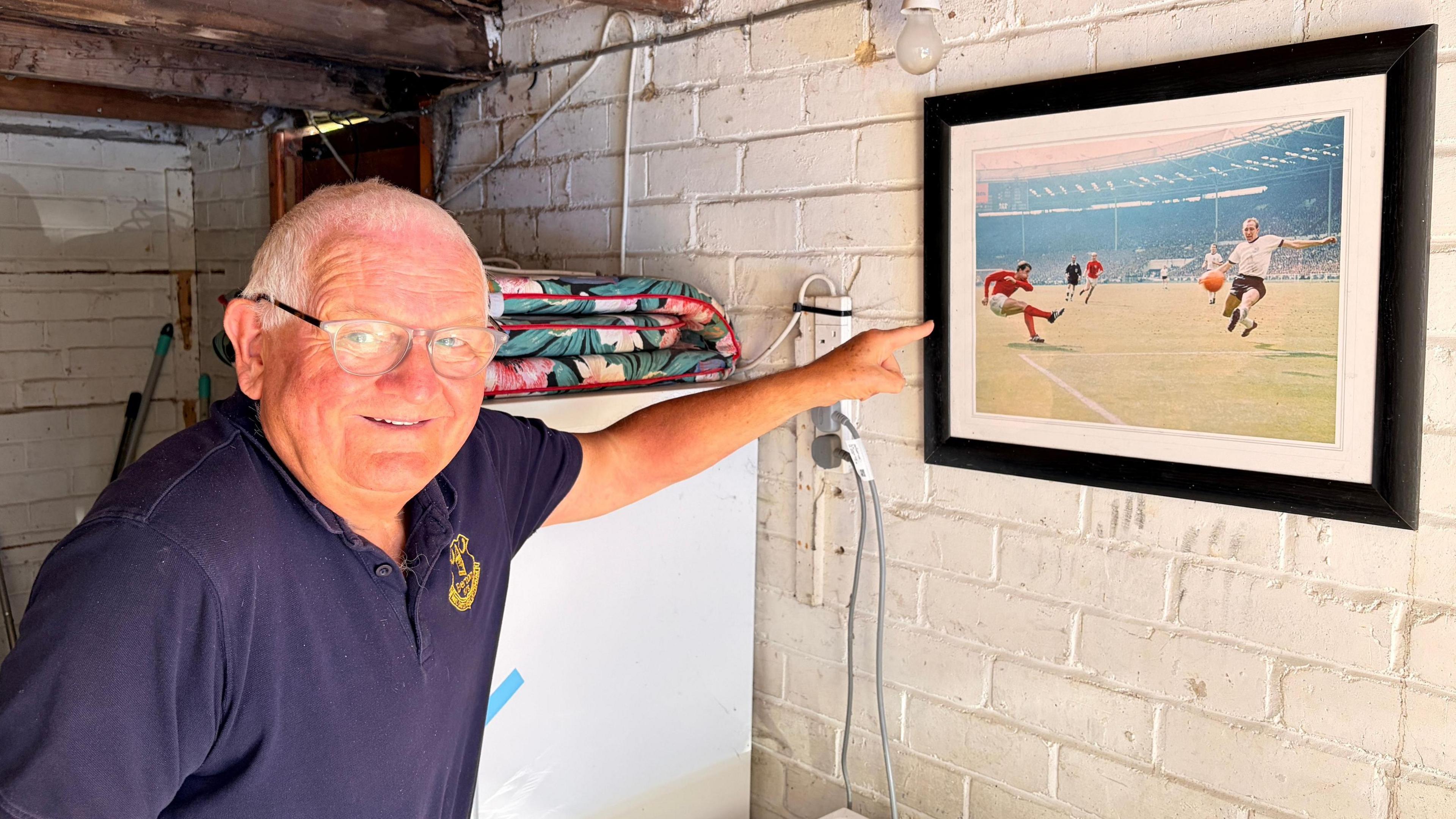 A man in a navy blue T-shirt points at a picture of the 1966 World Cup game hung on a wall. The man is smiling.